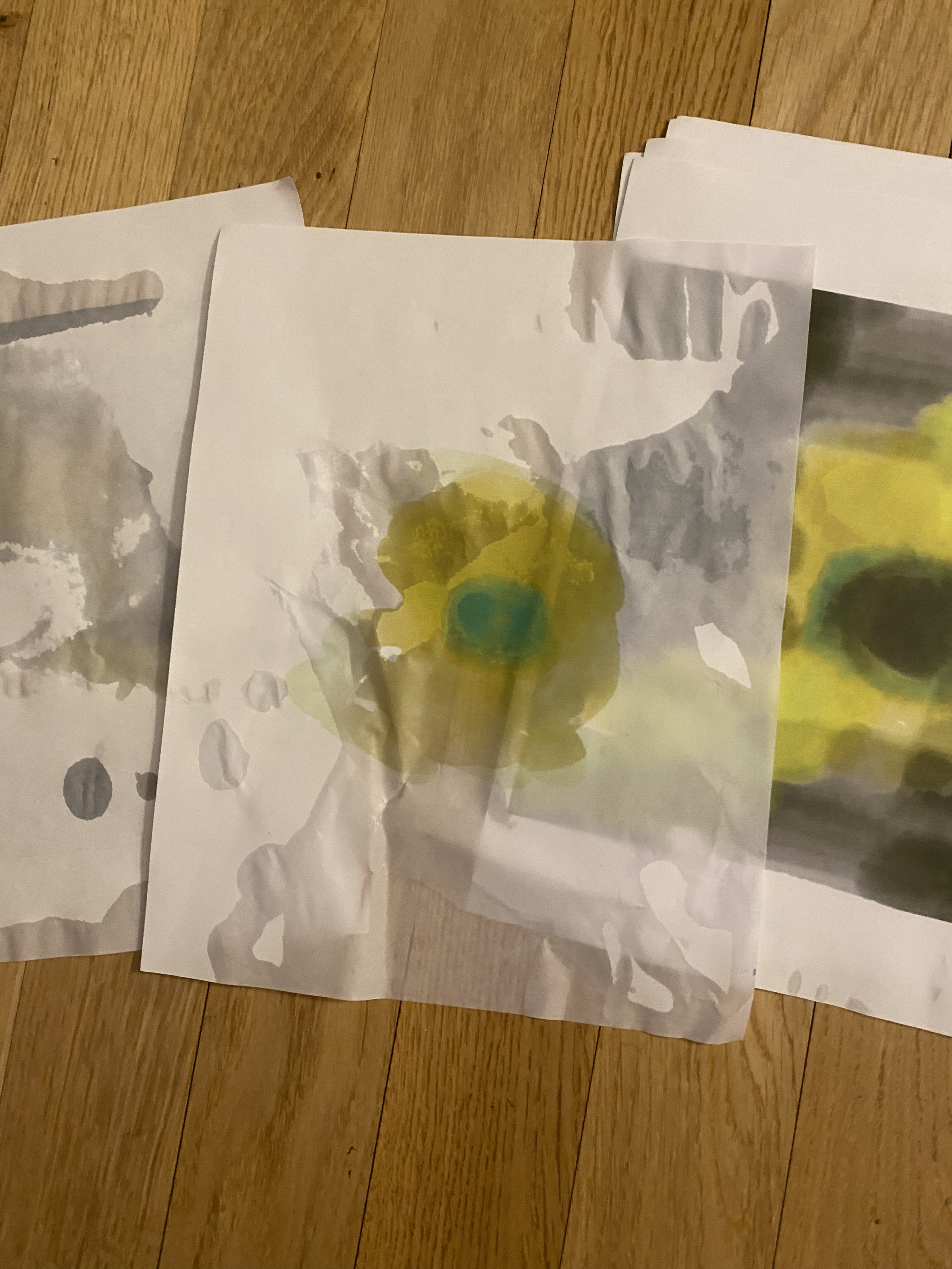 Several pages of a yellow and green abstract painting is laid out on a wooden floor. Water has been spilled over them.
