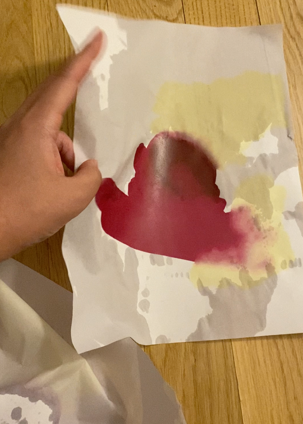 A hand holds a wet piece of paper with the print of a yellow, dark red, and magenta abstract painting.