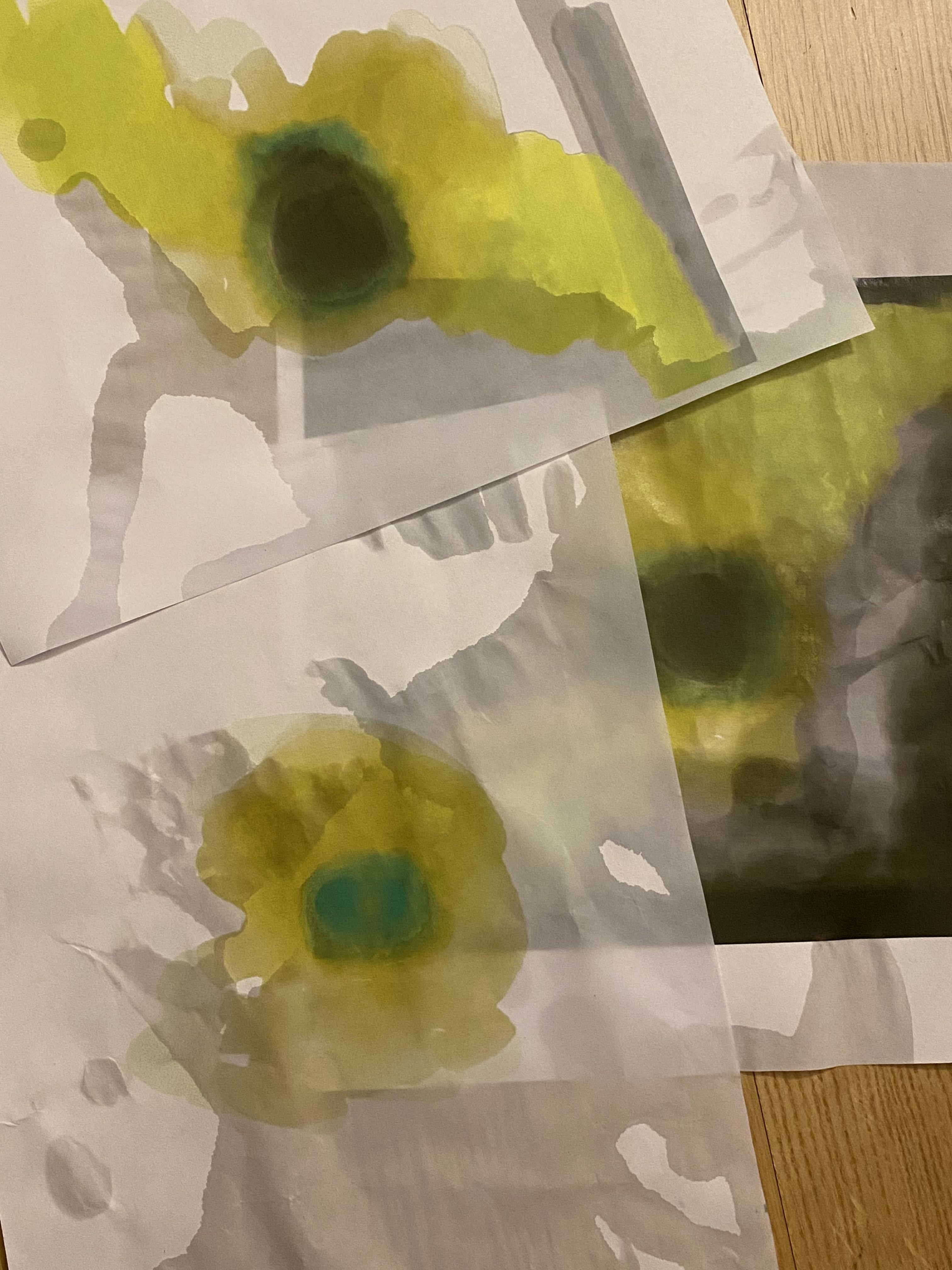 Several pages of a yellow and green abstract painting is laid out on a wooden floor. Water has been spilled over them.