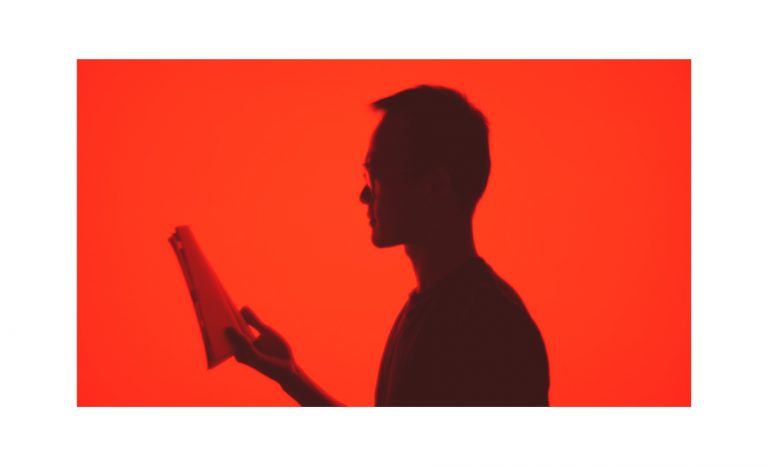 A photo of a silhouette of a man with glasses holding and reading a book. The background is red. 
