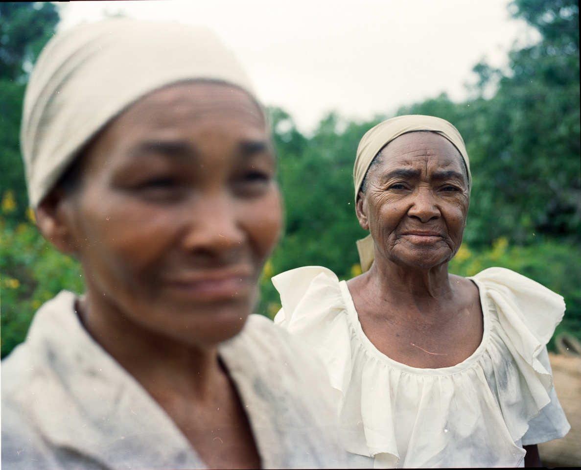 Image Description: A photo of two older women wearing light tan clothing and head coverings. The camera focuses on one woman in the foreground who looks directly at it, while the woman closest to the lens is out of focus.