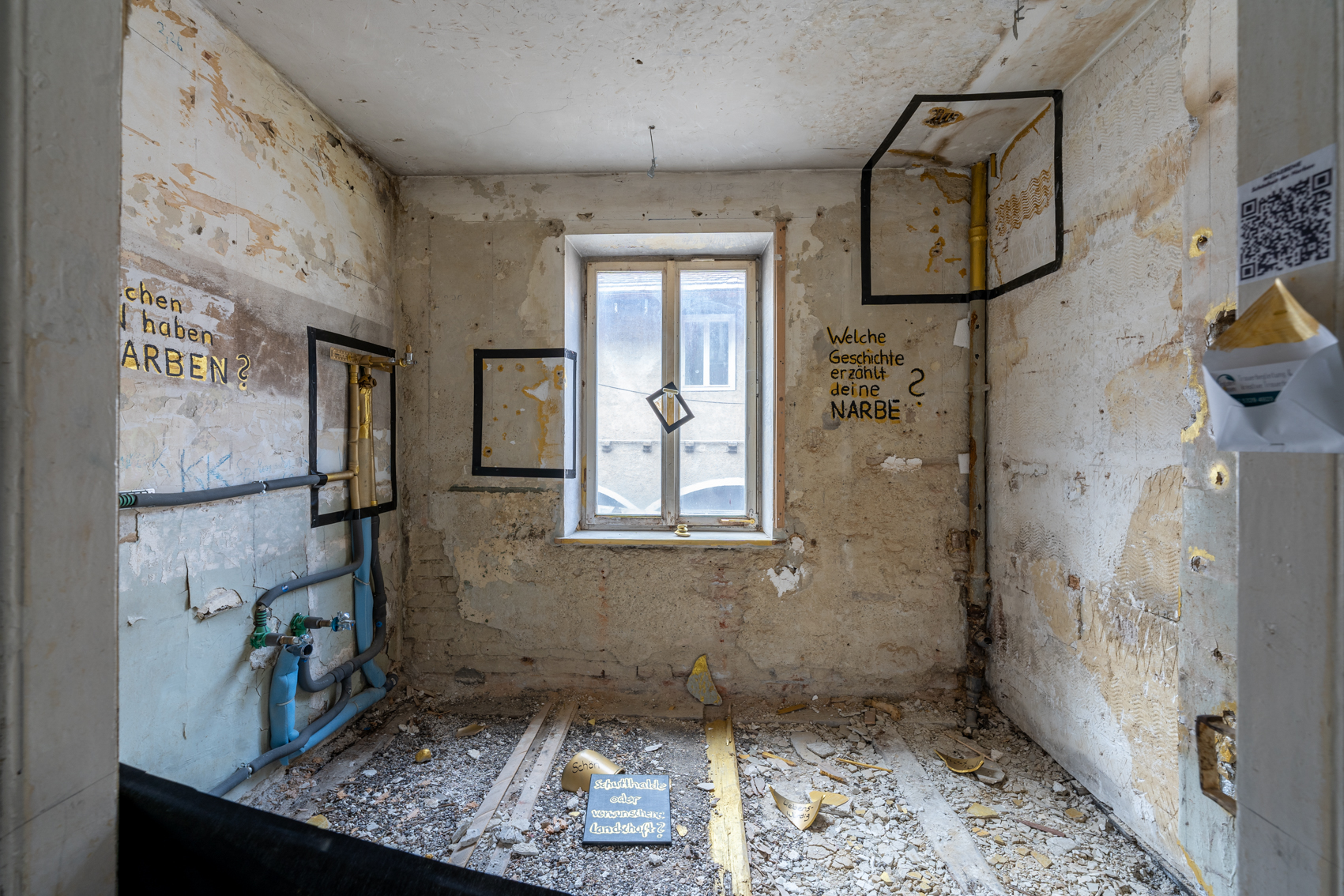 Bild 1 zu Sch&ouml;nheit der Narben: Blick in unverputzten Raum mit Schutt am Boden. An der gegen&uuml;berliegenden Wand befindet sich ein Fenster. Im rechten und linken Eck verlaufen Rohre. Teile im Raum wurden golden bemalt. Auf dem Boden steht auf einer kleinen Tafel: "Schutthalde oder verwunschene Landschaft?" An der Wand steht mit Handschrift: Welche Geschichte erz&auml;hlt deine Narbe?