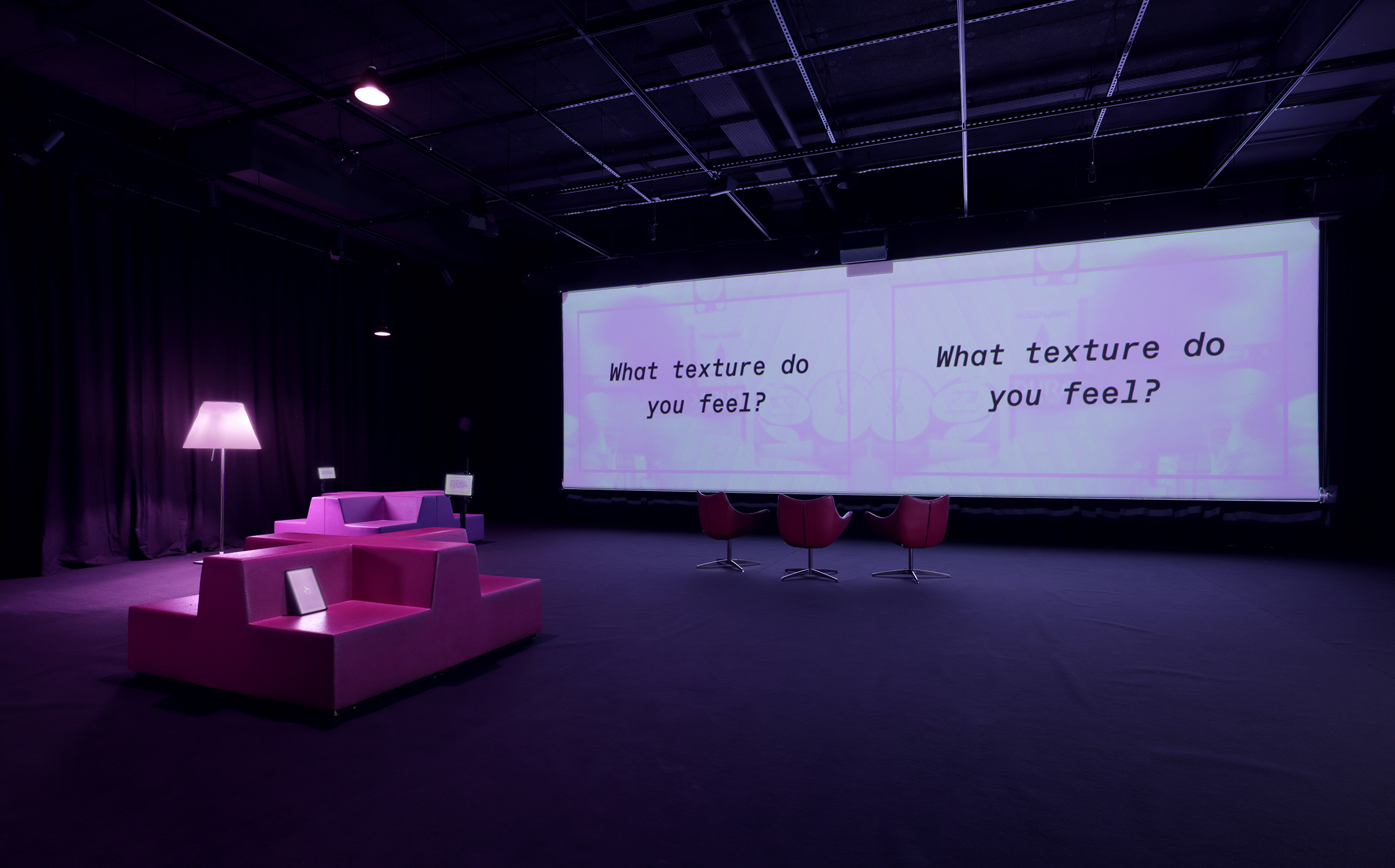 dark purple tinted installation with widescreen projection that says what texture do you feel, box-like seating and purple tablets