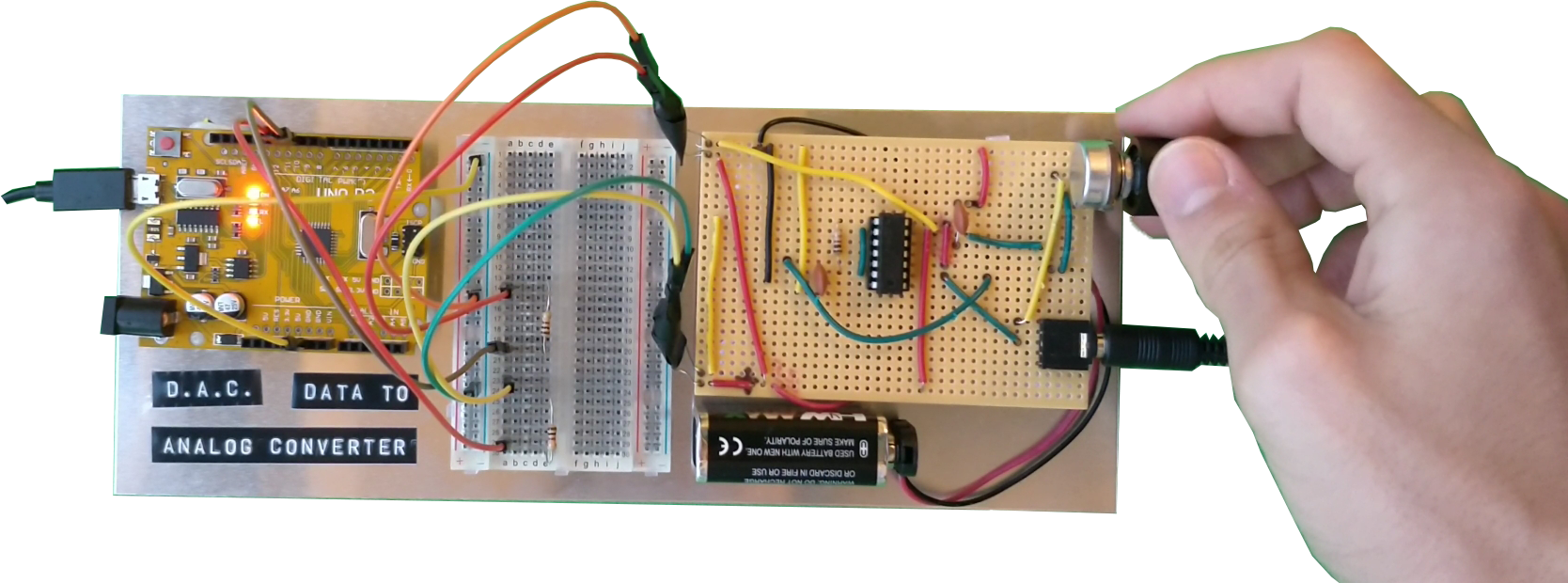 An array of circuitry mounted on a small sheet of aluminium. Thin cables run between the circuit boards with a power cable plugged into the left hand side and an audio cable plugged into the right. A hand is turning a volume knob at the top left corner of the aluminium sheet.