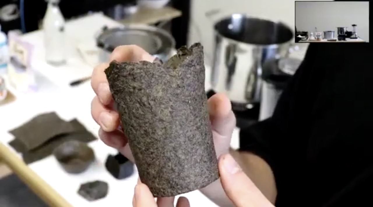 Person holding a vessel created from invasive plant material up to their zoom camera during a workshop. In the background there are other items created with invasive plant material, pots, bowls and other laboratory equipment.