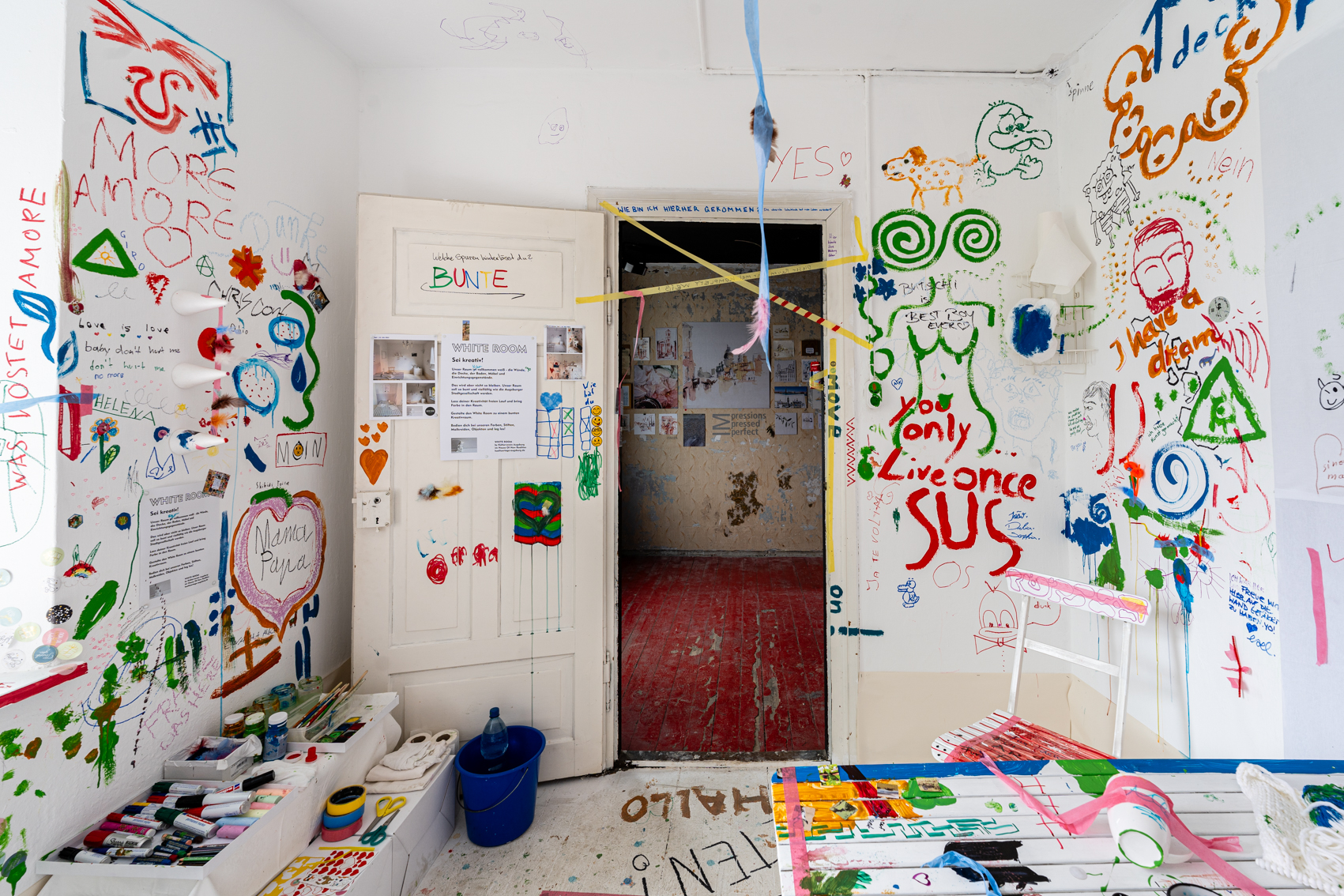 Bild 3 zu White Room: Im Raum stehend mit Blick auf die ge&ouml;ffnete wei&szlig;e T&uuml;r. Links der T&uuml;r  liegen Stifte, Pinsel und Farben.