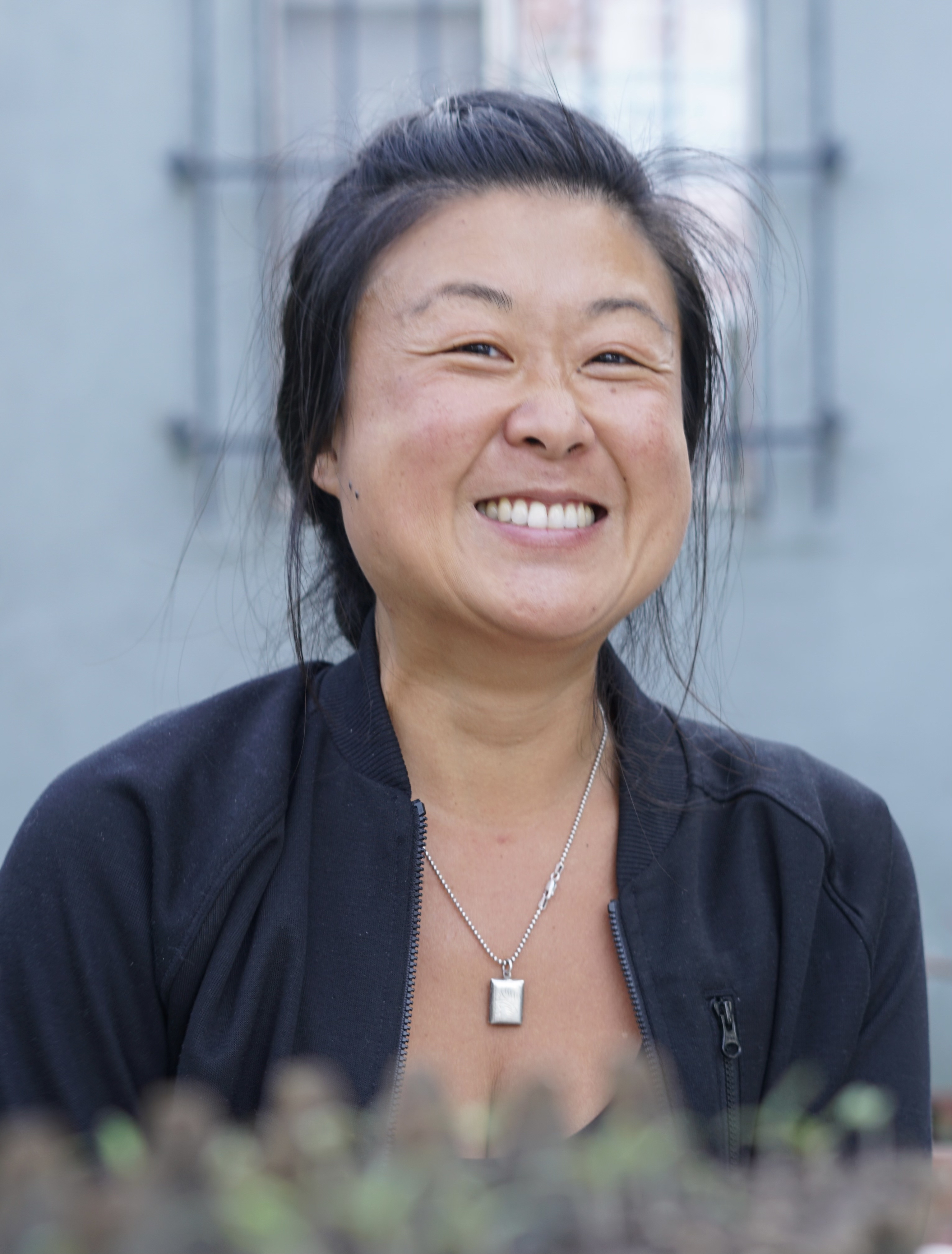 Photo shows Theresa Hyuna Hwang smiling in front of her home.  Photo credit: taken by her child with support from her partner David Malana