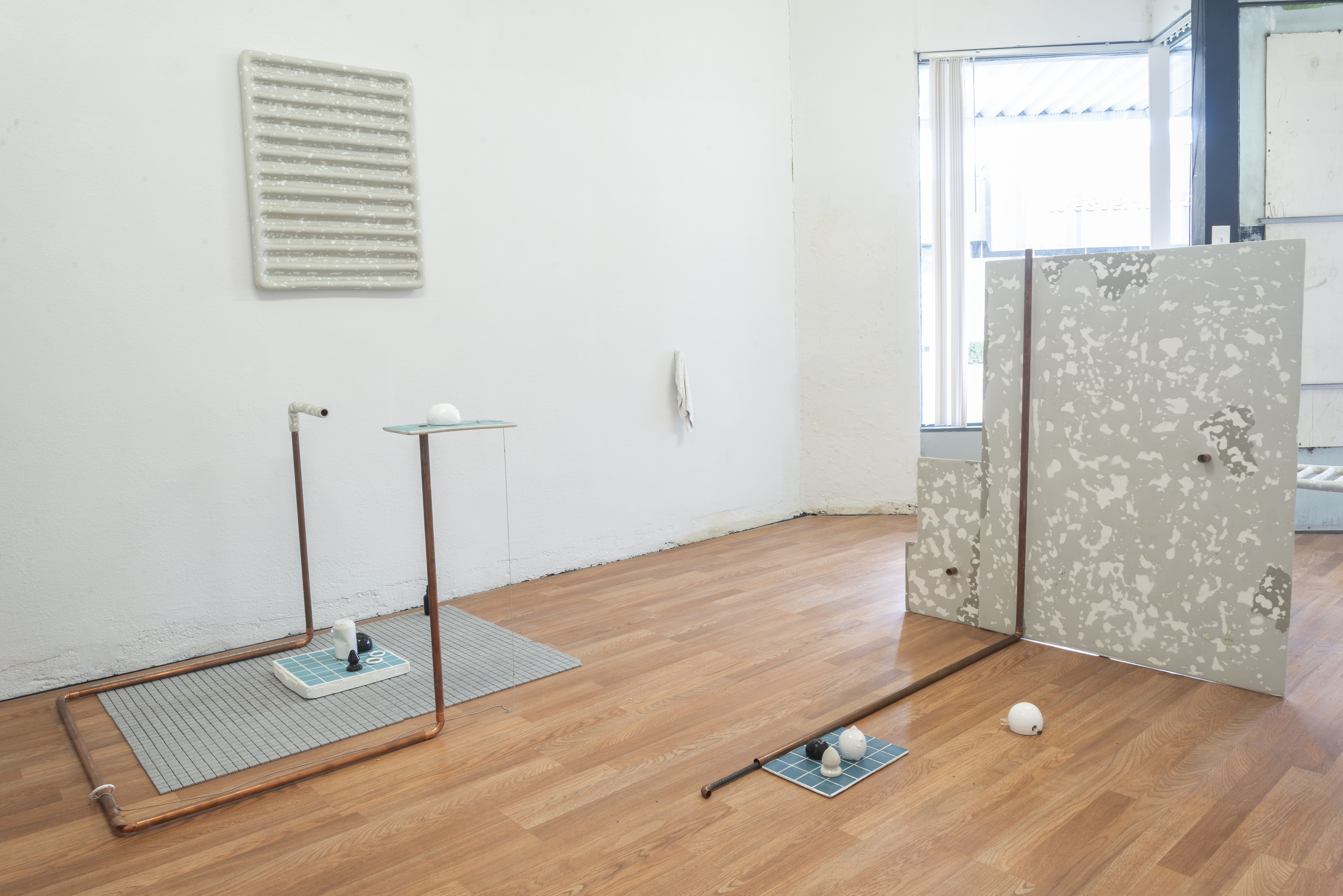 The same gallery space, but from behind certain sculptural objects revealing copper pipe, like the kind that might supply water to an older building. Distanced on the floor are smaller fragments of tiles and sculptures. Vignettes or snapshots of a scene.