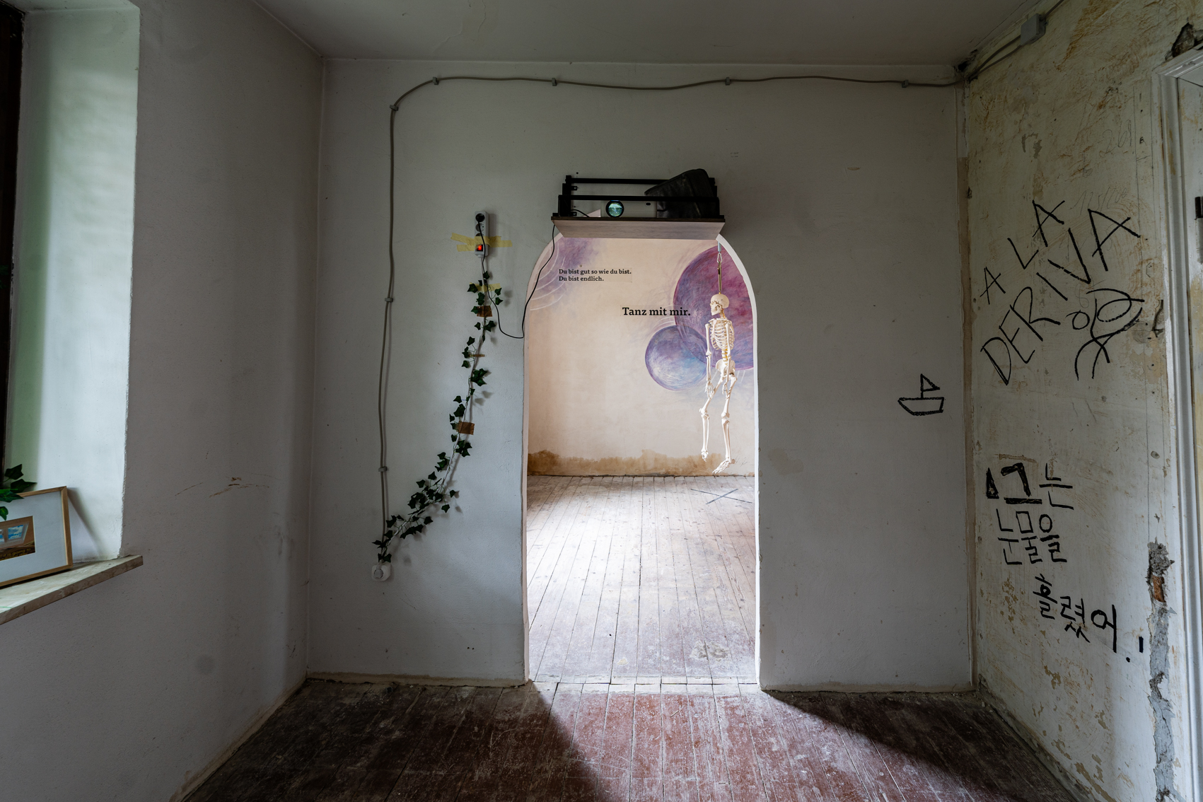 Bild 2 zu A la deriva: Blick im Raum stehend in Richtung Rundbogen zum Raum des Augsburger Totentanz. Oberhalb des Durchgangs befindet sich der Beamer. An der Wand rechts steht in schwarzer Schrift "A la deriva". Darunter befinden sich handgemalte schwarze Zeichen. 