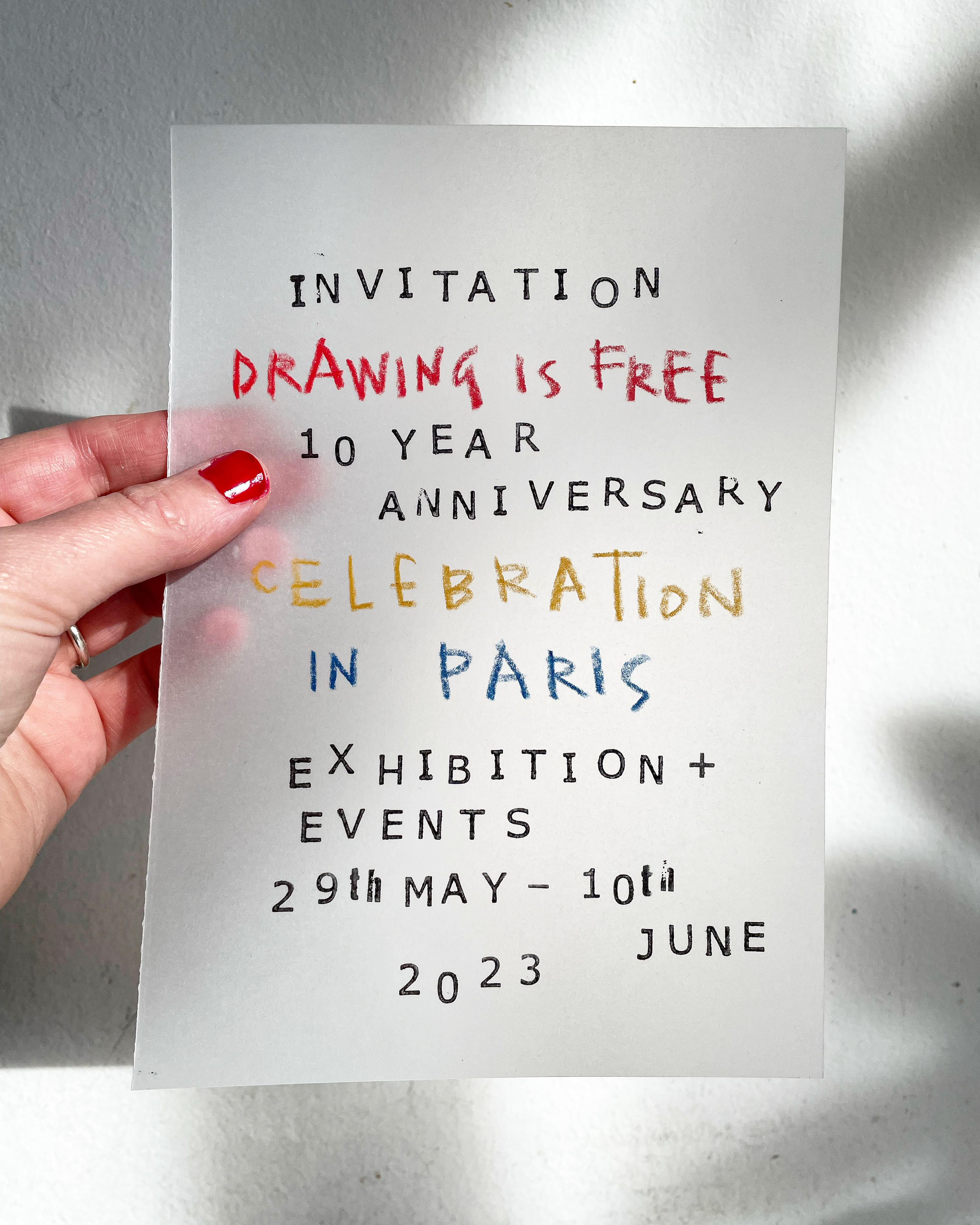A hand holding a hand lettered invitation to the Drawing is Free 10 Year Anniversary Celebration in Paris 29 May-10 June 2023