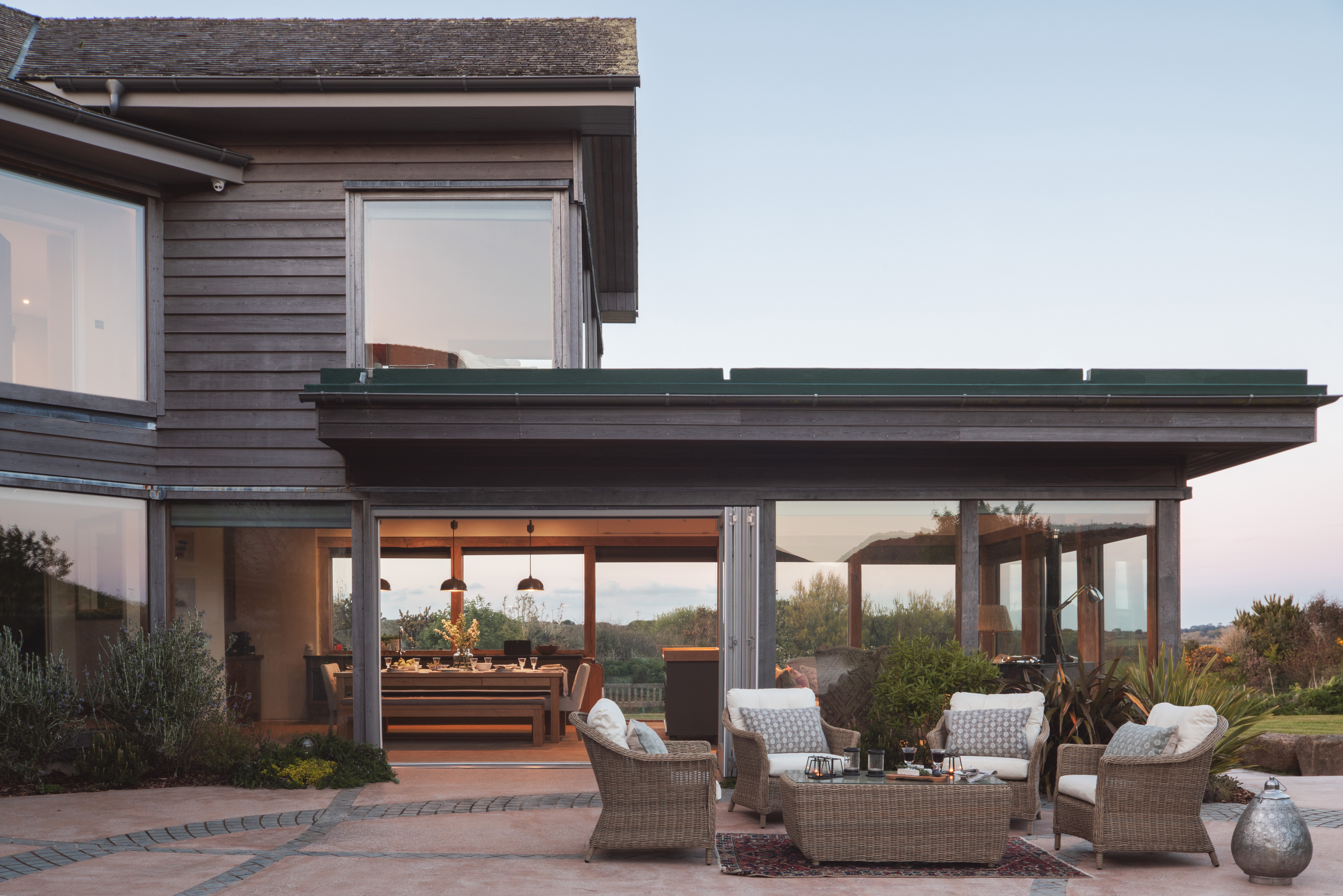 Exterior of residential home at dusk. Facade is clad in grey timber. Paved patio with rattan outdoor seating and table.