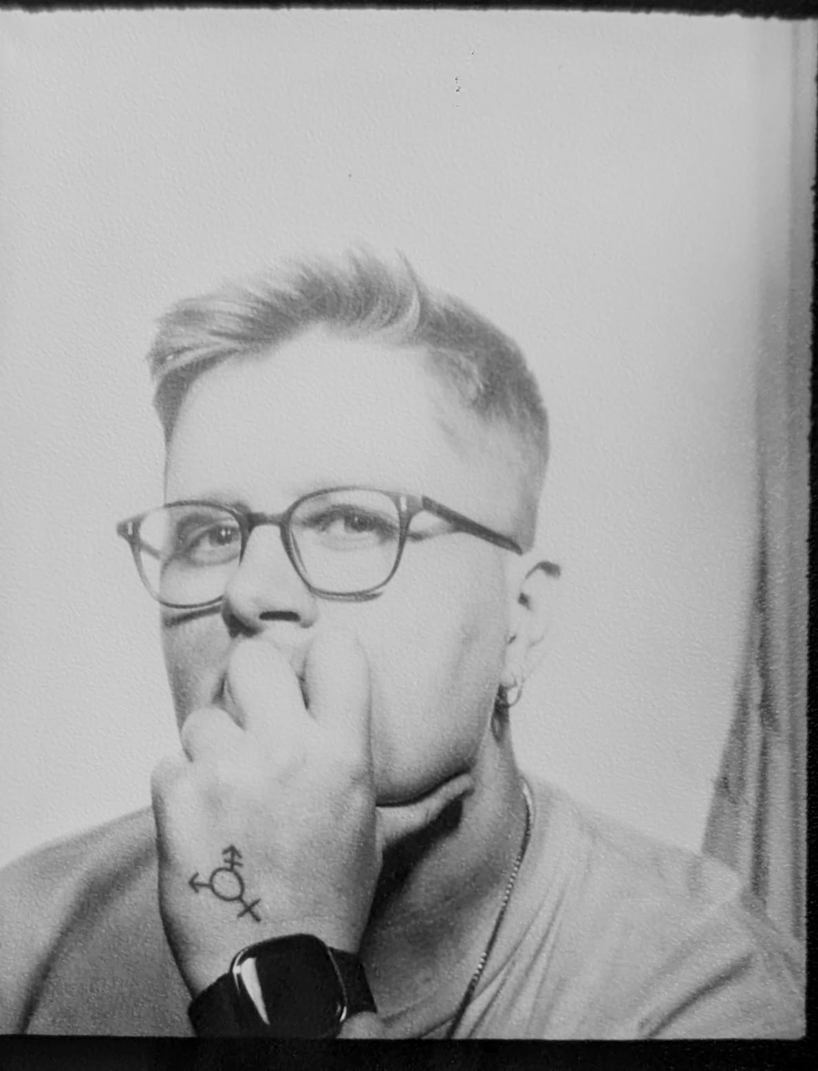A black and white Photo Booth portrait of Edi, who is a white, butch person with short back and sides hair to a skin fade and is wearing glasses. They are holding their chin and looking at the camera, with a tattoo visible on their hand of the transgender symbol.
