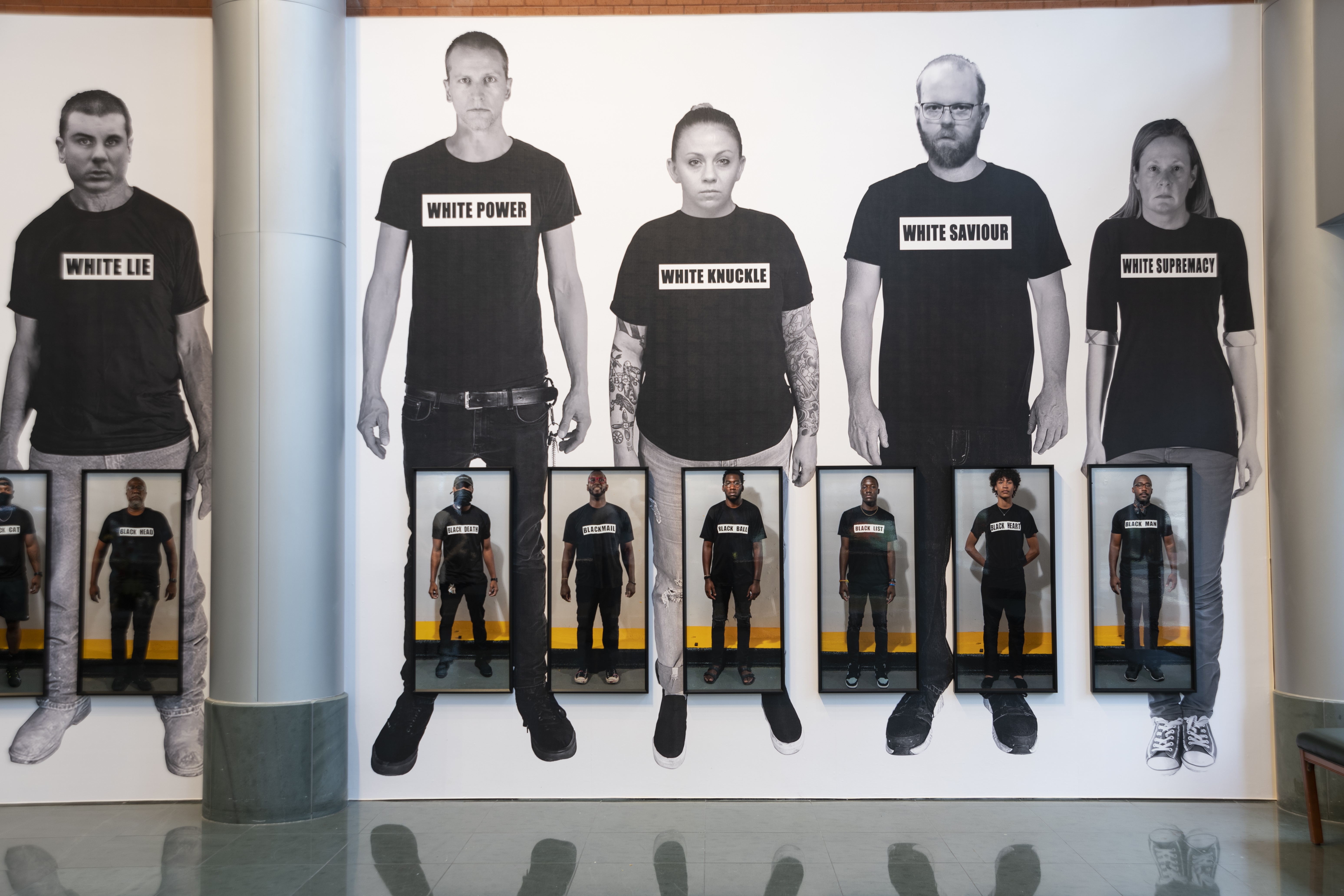 A white gallery wall displays a larger-than-life lineup of figures with lighter skin tones, each wearing black shirts while a lineup of framed, smaller, life-sized photographs of figures with darker skin tones are hung overtop in a line closer to the floor.