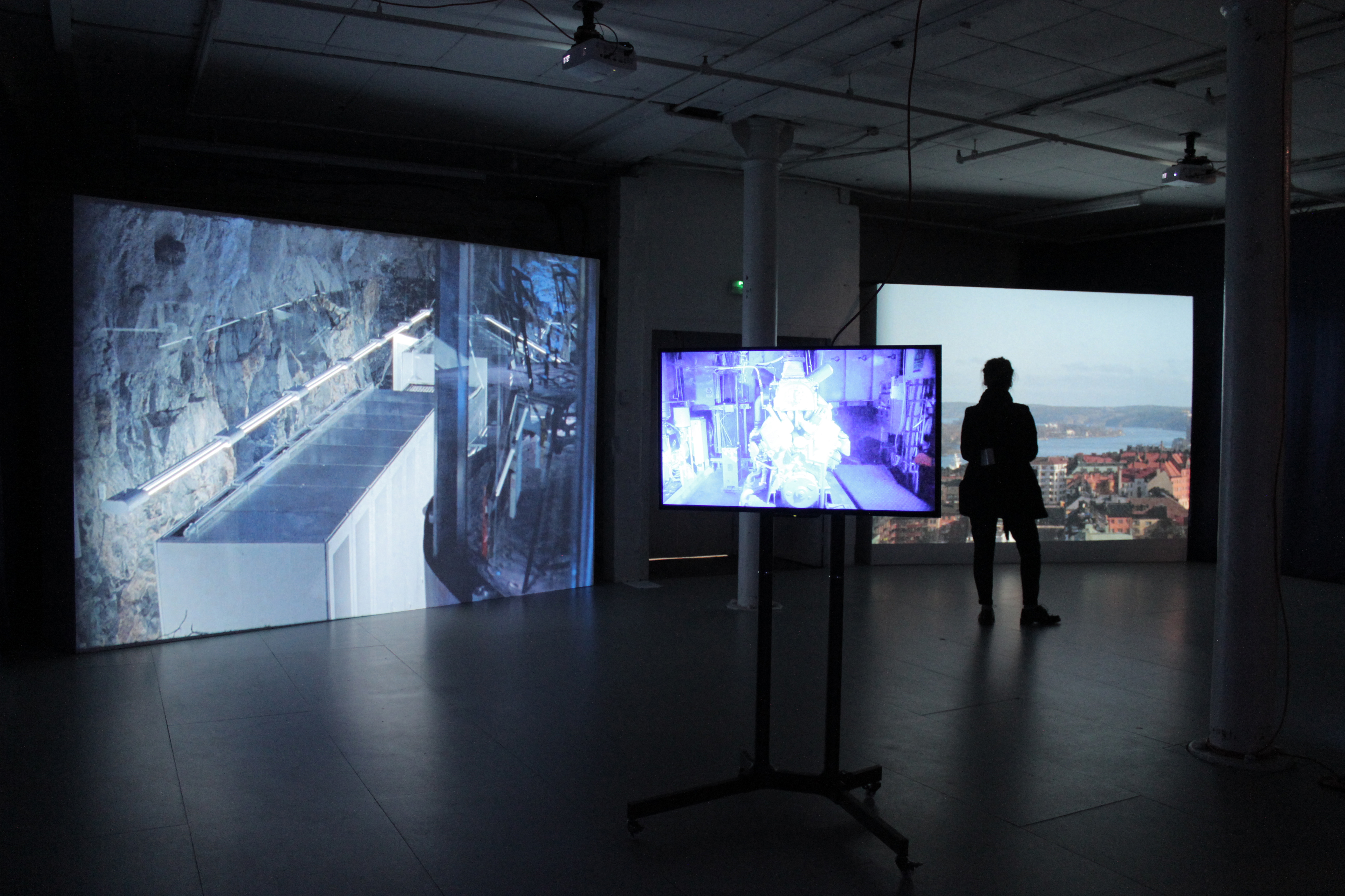 There is a projected video to the left, a flatscreen tv on a stand in the middle and a second projection on the right side of the frame. On the right side is also a person facing the screen.