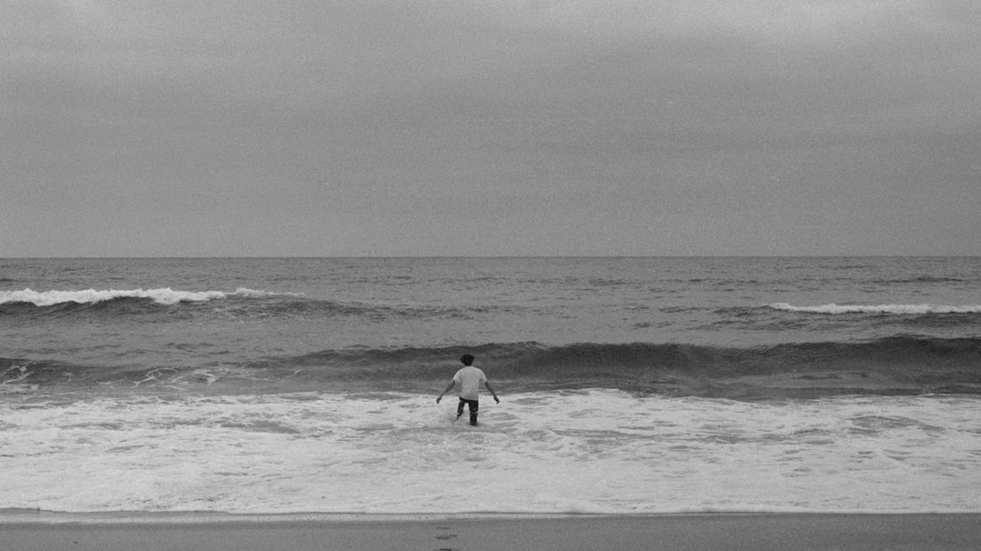 ID: Black and white image of a man in a white shirt wading into the ocean. The man is in middle-distance, and the sky is overcast.