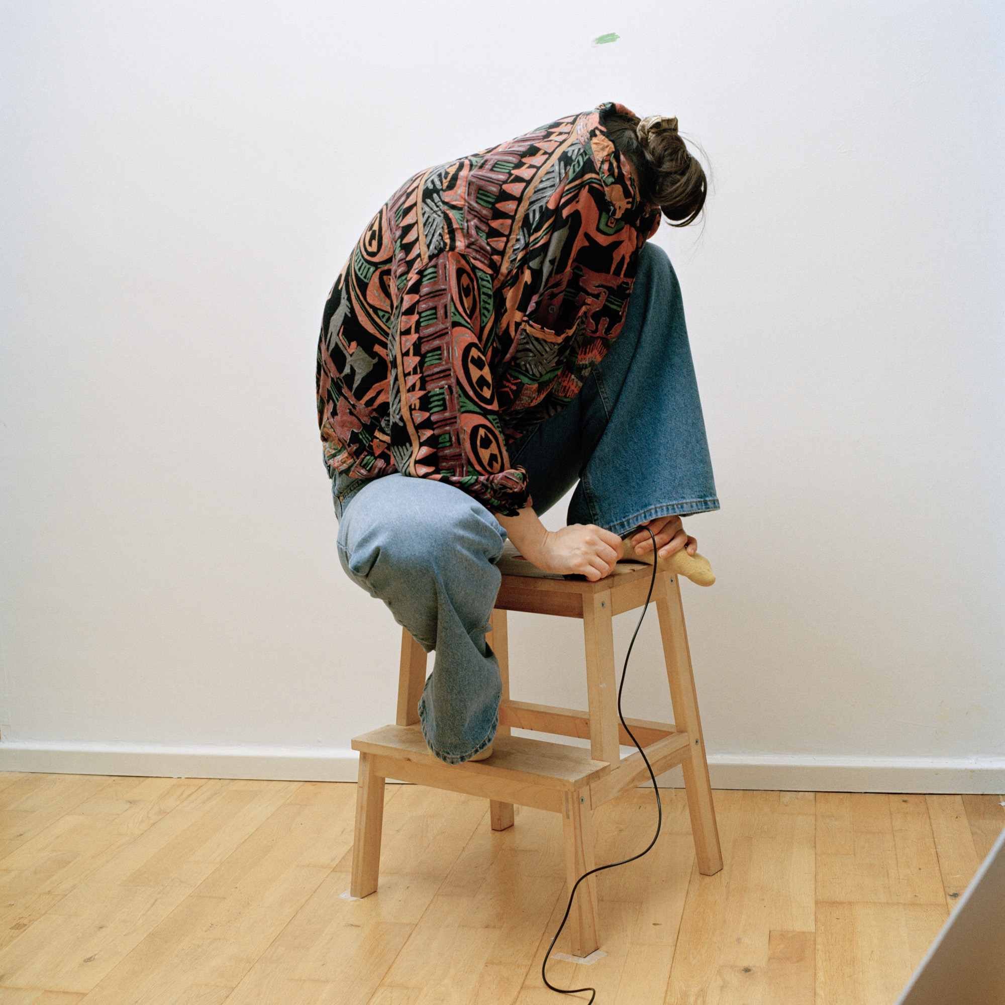Performative Self Portrait of the artist, sitting crouched on a chair face hidden by their clothes and hand holding a shutter release shot by UK-based photographer Ilayda Akarca