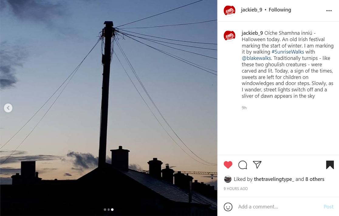 Instagram post by @Jackieb_9 showing the tips of rooftops with a power line reaching into the sky and a blue sky with orange and yellow at the bottom. The text reads: Oíche Shamhna inniú - Halloween today. An old Irish festival marking the start of winter. I am marking it by walking #SunriseWalks with @blakewalks. Traditionally turnips - like these two ghoulish creatures - were carved and lit. Today, a sign of the times, sweets are left for children on windowledges and door steps. Slowly, as I wander, street lights switch off and a sliver of dawn appears in the sky.