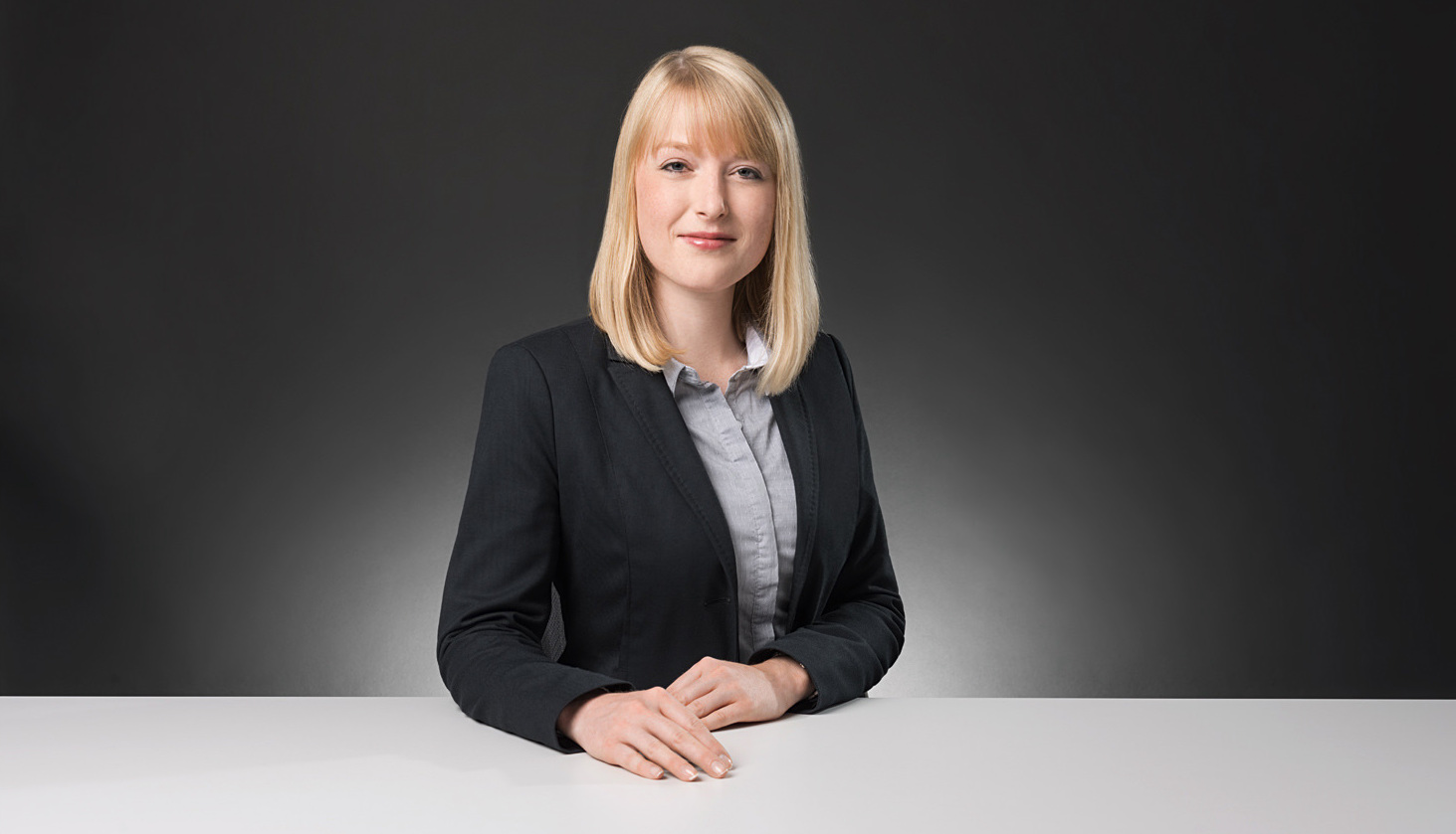 Business-Portrait einer Geschäftsfrau, sitzend vor grauem Hintergrund.