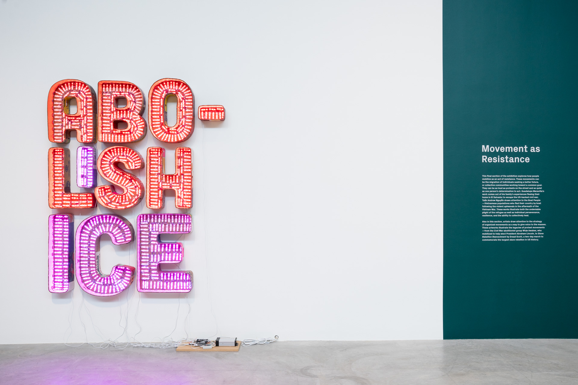 A sculpture on a white wall made to look like flashy light-up sign lettering, as if for a business, that spells out “Abolish Ice” in three lines, with “ABO,” and “LISH” taking up the first two in a red light and “ICE” taking up the bottom line in purple. Small wires fall out of this to the floor where there is some kind of electronic controller. To the right is a section of the wall painted a blue-green with white vinyl wall text for the show title at the top, “Stories of Resistance.”