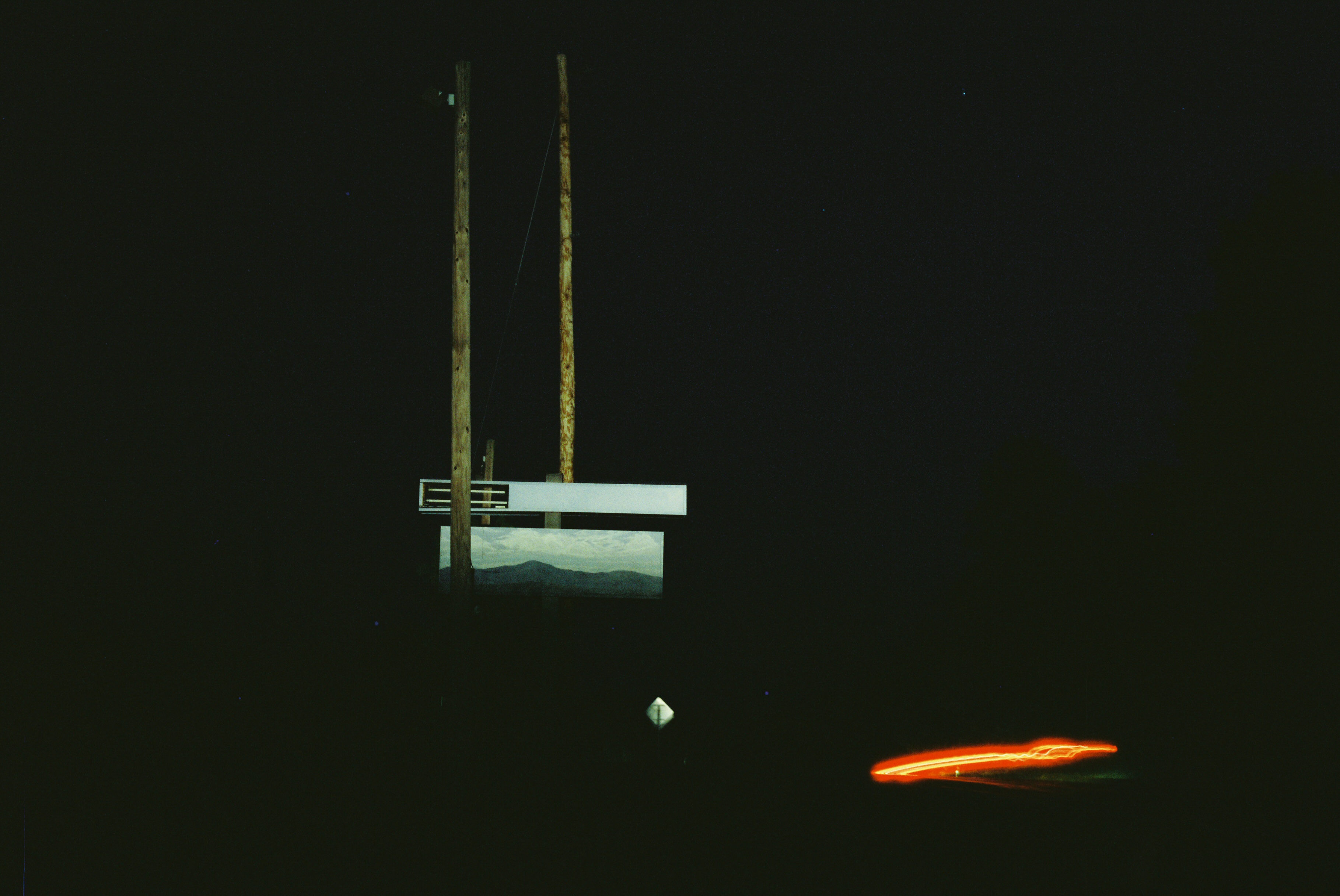 Delmar Coffin BuildingImage Description: The image is pitch black, aside from two main areas. In the bottom right corner, there is a vaguely horizontal red neon trail, like the tail lights of a moving car. Slightly left of center, coming from near the top of the image down, two wooden poles are visible, appearing parallel but with the left more in the foreground than the right. A sliver of wire moves from the right pole diagonally to the left. Just below the center of the image, there are two rectangles that are intersected by the left pole. The upper rectangle is a thin metal slat, the left end of which looks like a grille. The lower, which is the larger of the two, is a painting of a mountain against a pale blue-and-green cloudy sky. It looks like it's a billboard, and it is positioned above the illuminated back of a diamond-shaped street sign.