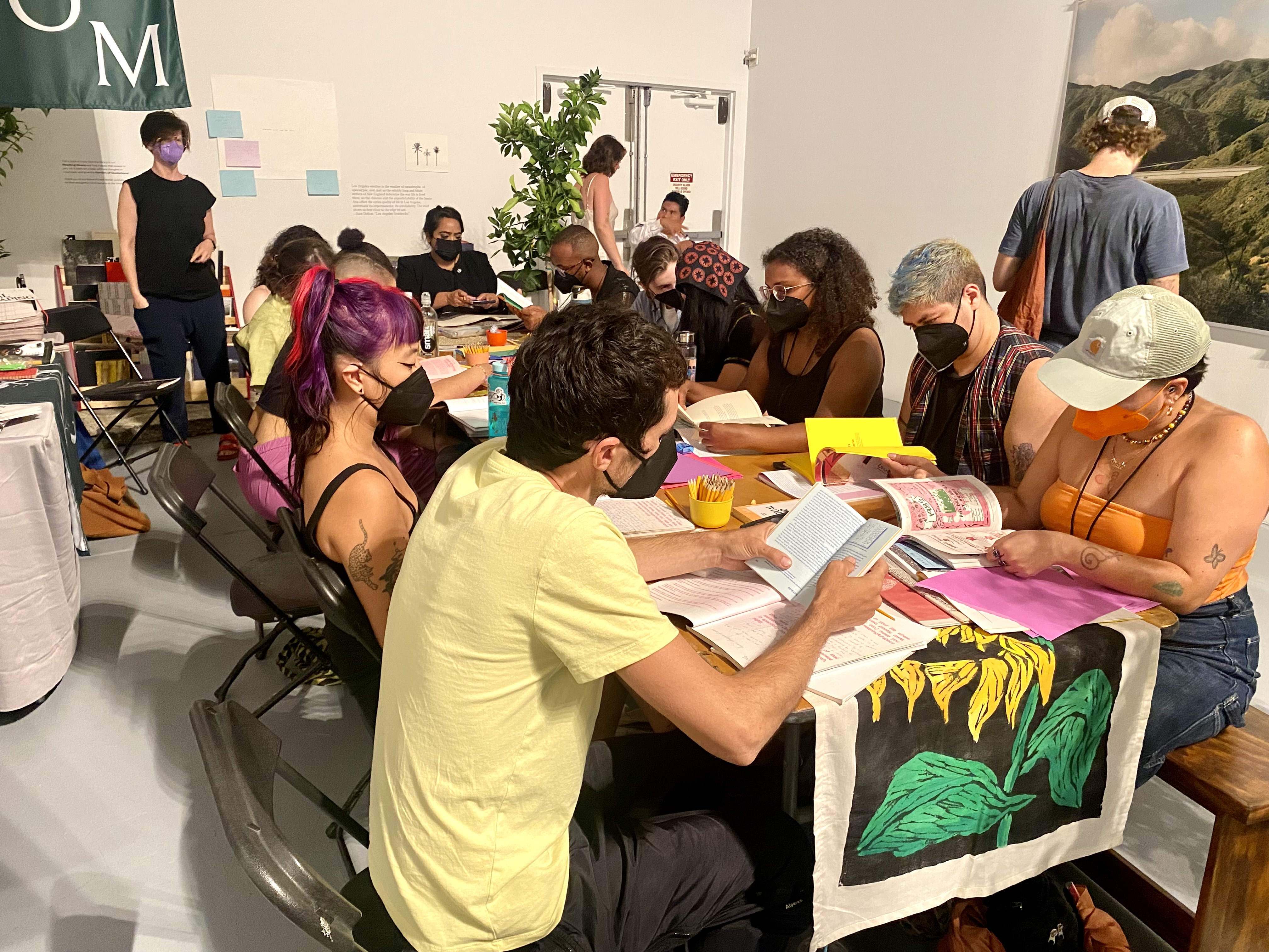 A collection of masked people gather around a long table paging through zines and political flyers.