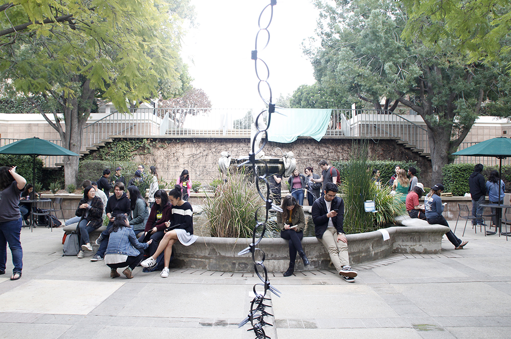 View of a courtyard where many people sit and hang out on the edge of a fountain that is filled with aquatic plants. A thin chain-looking artwork goes across the view vertically in the center.