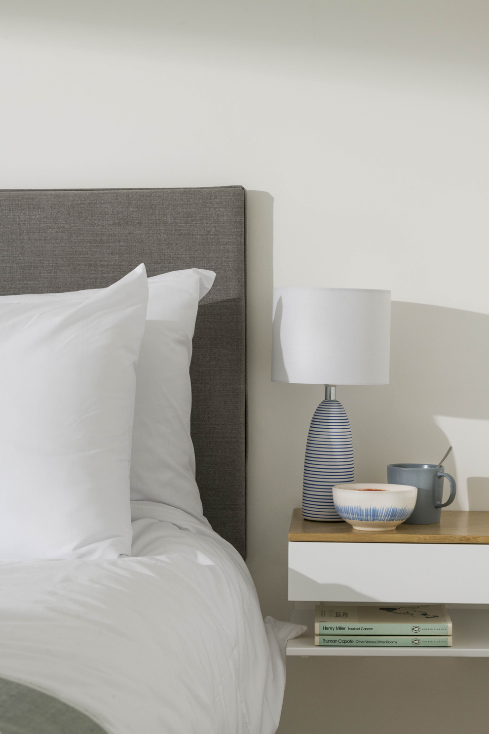 Close-up of upholstered bed headboard and side table with lamp, ceramic tableware and two books. 
