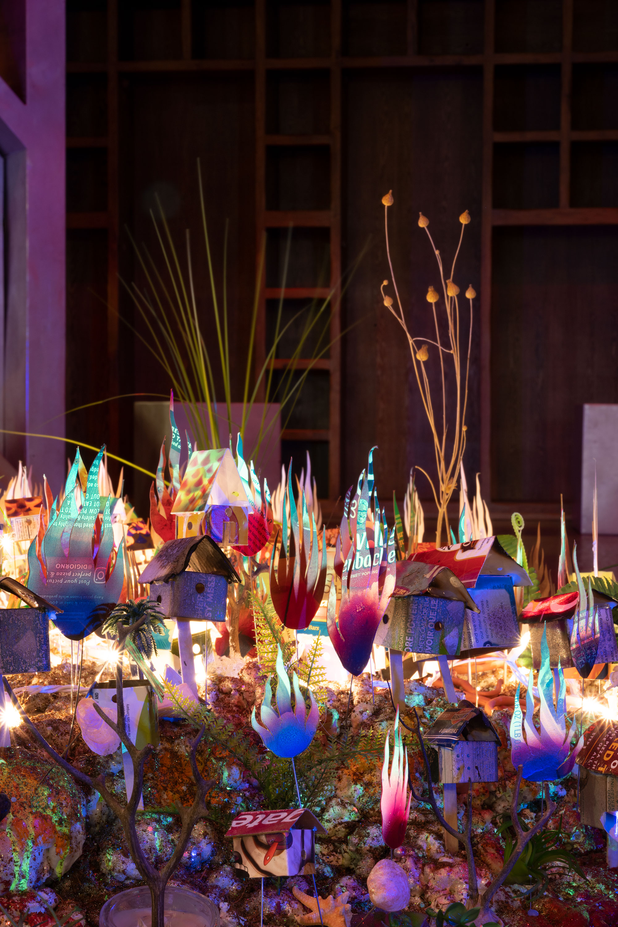 Orlando Estrada, Tiered Panorama, (detail) 2021, coconut fiber, expanding foam, glitter, artificial pearls, 3D printed PLA filament, sea shells, stone, artificial foliage, cell phones, Swarovski crystal, bone, consumer waste, found objects, light string, LED light, motor, UV pigment, acrylic, adhesive, PVC and Wire Armature 60” x 120” x 120”. Illuminated by strings of lights within the foreground, miniature houses made from consumer waste stand on stilts alongside various plants and objects as bushels of fire surround them.