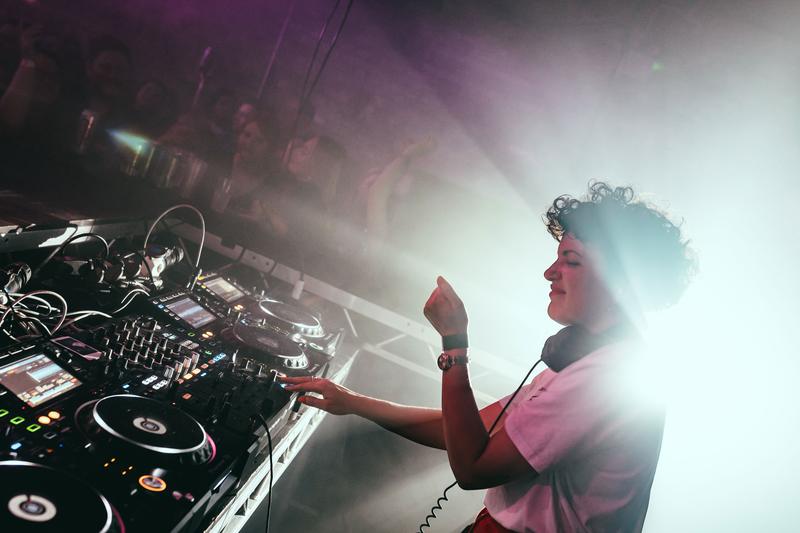 Annie Mac performing at floor-level DJ decks in Banana Block, with bright beams cutting through the Belfast crowd.