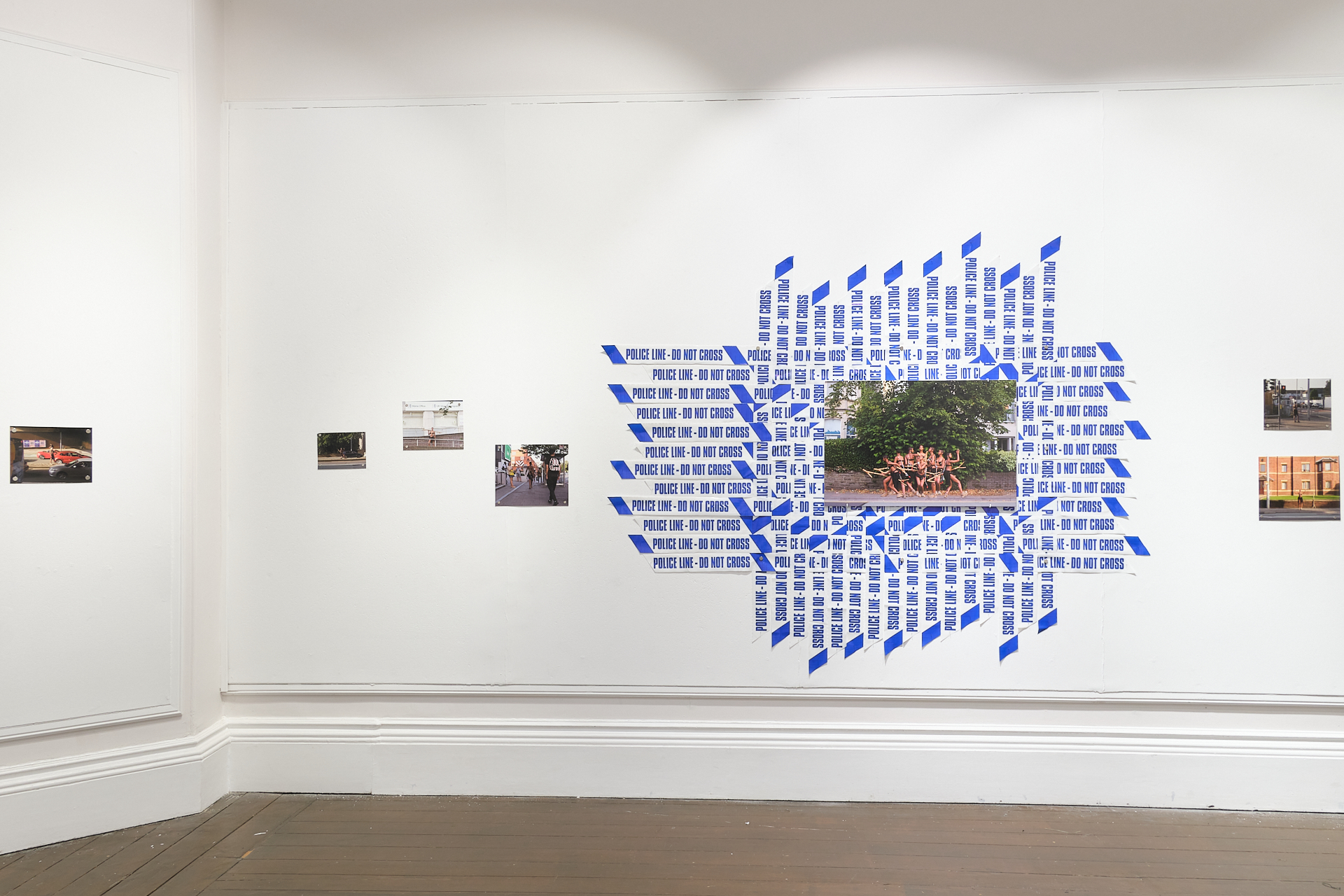 Set across the backdrop of a sunny street with terrace housing, six uniform figures of Aisha in a black skirt carry a branch - five figures levitate, one stands on the pavement, all facing away from the viewer