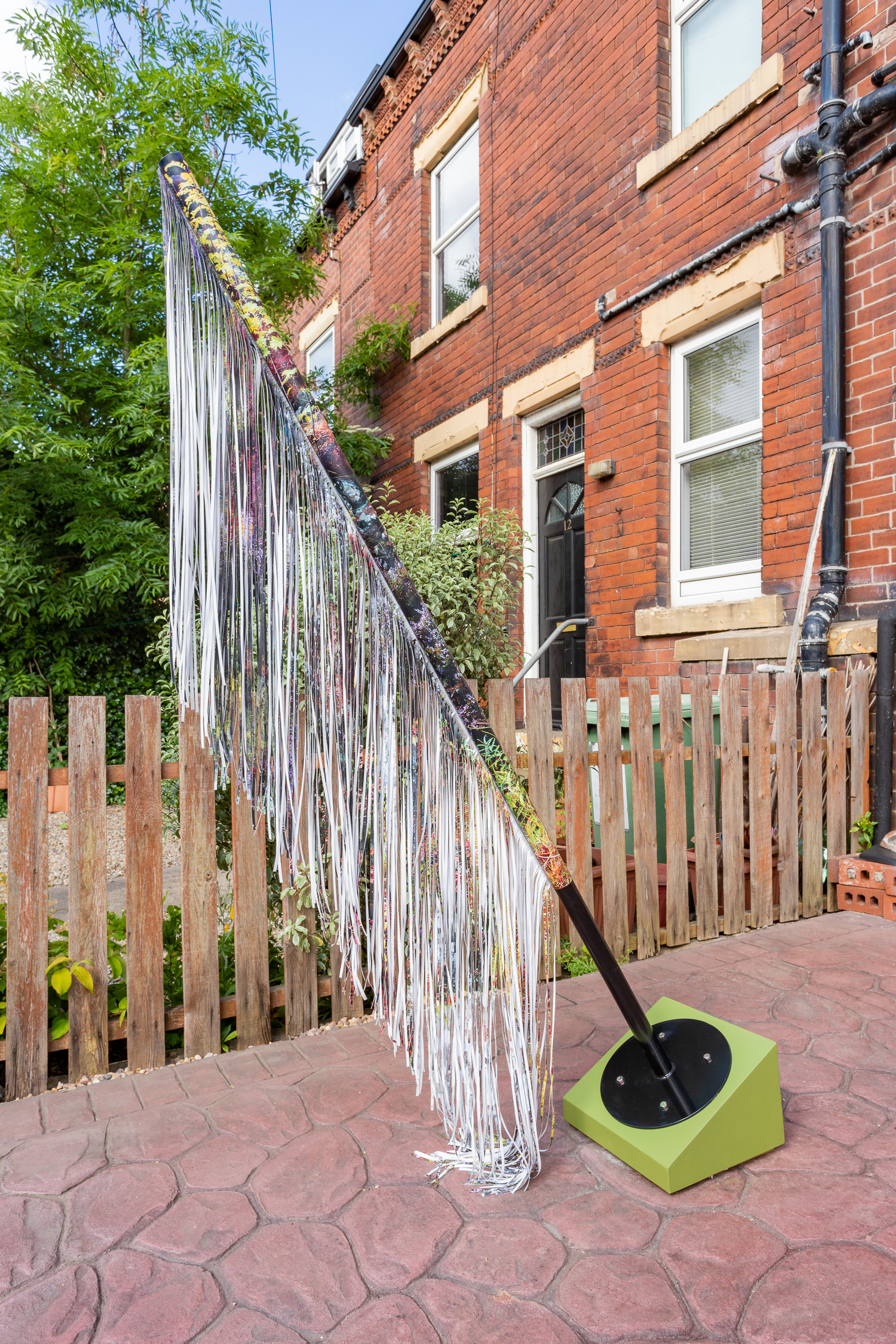 The Strata of Things I, 2021. A black steel stand on an angle sits on a green plinth, a photograph on vinyl is wrapped around the pole, with most of the image cut into thin strips like a fringe that falls to he floor.