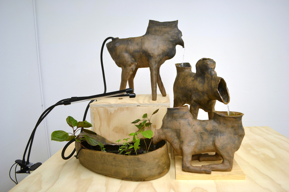 A series of brown ceramic vessels that resemble 4-legged animals without heads, which are acting as a kind of fountain, with the help of a small electric pump. Water flows between them.