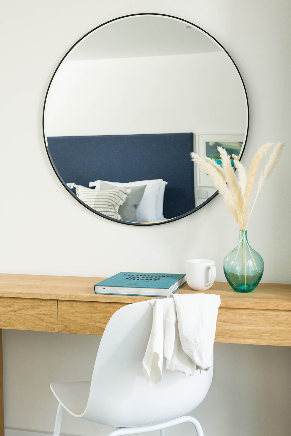 Close-up of wooden desk with blue glass vase, cup, book and white chair. Wall mounted round mirror.