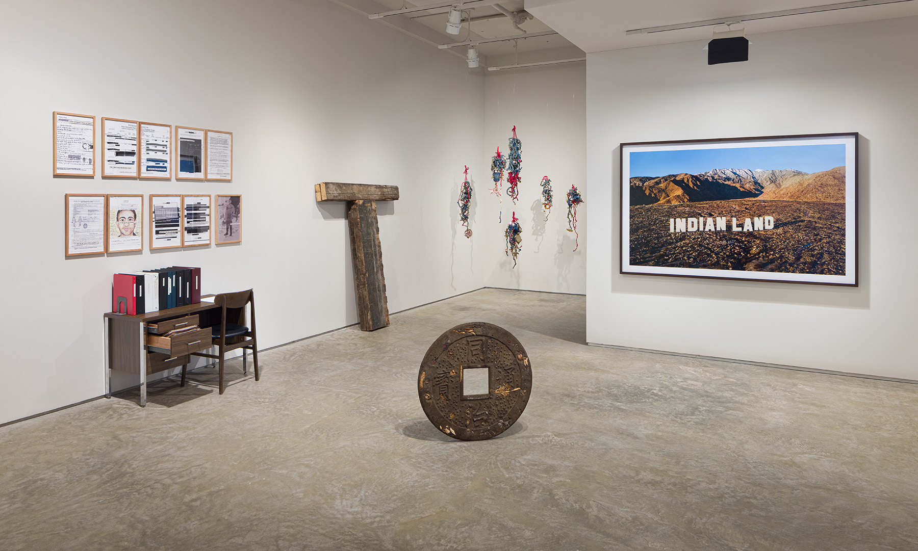 An Incomplete Haunting, Installation view. From left to right:Chitra Ganesh + Mariam Ghani, Introduction to an Index, 2011. Ten prints on Hahnemühle photo rag paper, binders, desk, chair. Prints: 11 x 17 inches each.Courtesy of the artists. Kris Grey, Capital T, 2025. Salvaged wooden joists from Stonewall National Monument Visitor Center, 75 x 60 x 6 inches. Courtesy of the artist. Elvira Clayton, Indigo Bess and her Children, 436 project, 2024. Six sculptures: indigo-dyed osnaburg, cotton, wire, jute, sequin, mini glass bottles, cotton bolls, shells, twigs, screen-printed archival text and photographs, embroidery hoops, acrylic, handwoven, and crochet. Bess: 37 x 12 x 7 inches; Sam: 46 x 12 x 5 inches; Elizabeth: 37 x 7 x 5 inches; Sena: 29 x 10 x 7 inches; Affa: 31 x 7 x 4 inches; Hannah: 37 x 10 x 6 inches. Courtesy of the artist. Kenneth Tam, Why do you abuse me, 2022. Epoxy resin, dirt, sand, dried mushrooms, dried seaweed, dried bamboo shoots, dried jujube, dried goji berries, preserved apricots, sunﬂower seeds, dried roots, dried sweet potato, steel, 30 x 30 x 1 1/2 inches. Courtesy of the artist and Commonwealth and Council. Nicholas Galanin, Never Forget, 2021. C-print mounted on Dibond, walnut frame, 51 3/4 x 78 3/4 inches. Courtesy the artist and Peter Blum Gallery, New York. Photo by Etienne Frossard.
