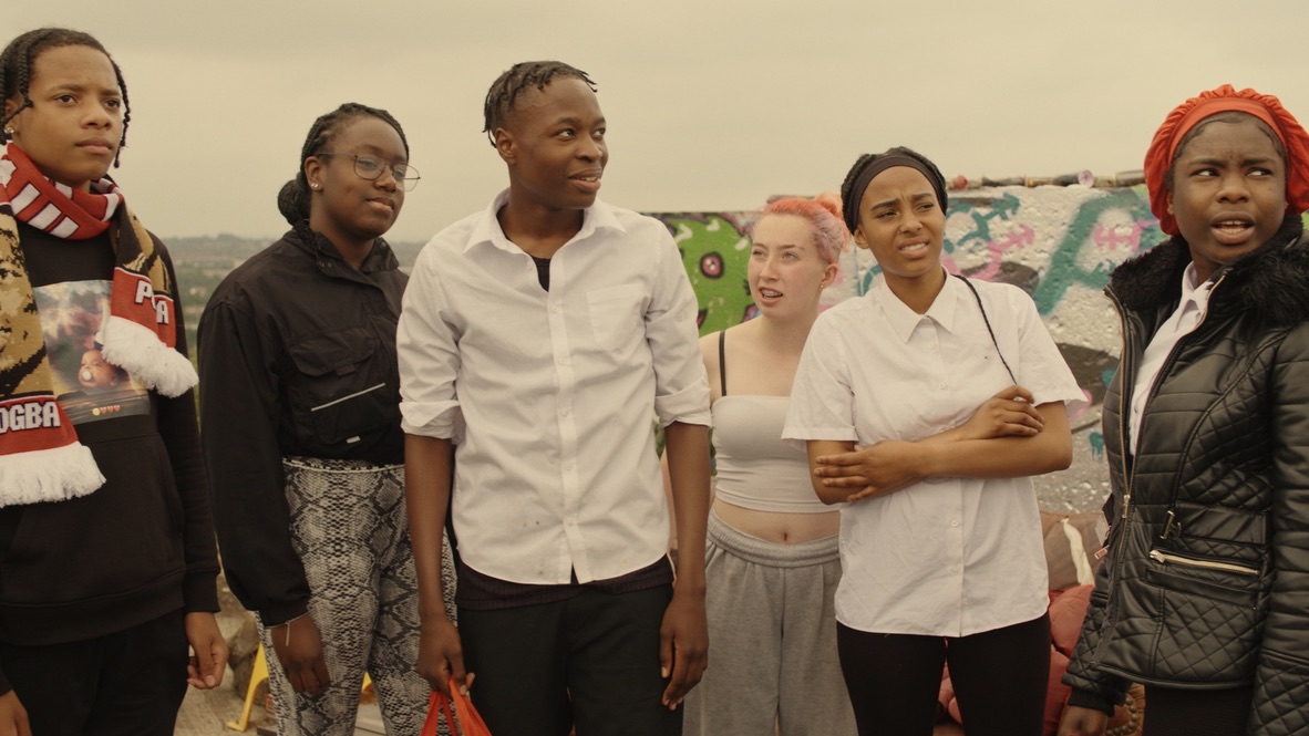 Son of Helios still. A predominantly Black group of young people stand outdoors wearing clothes that suggest they've just finished school. They look into the distance in mild indignation.