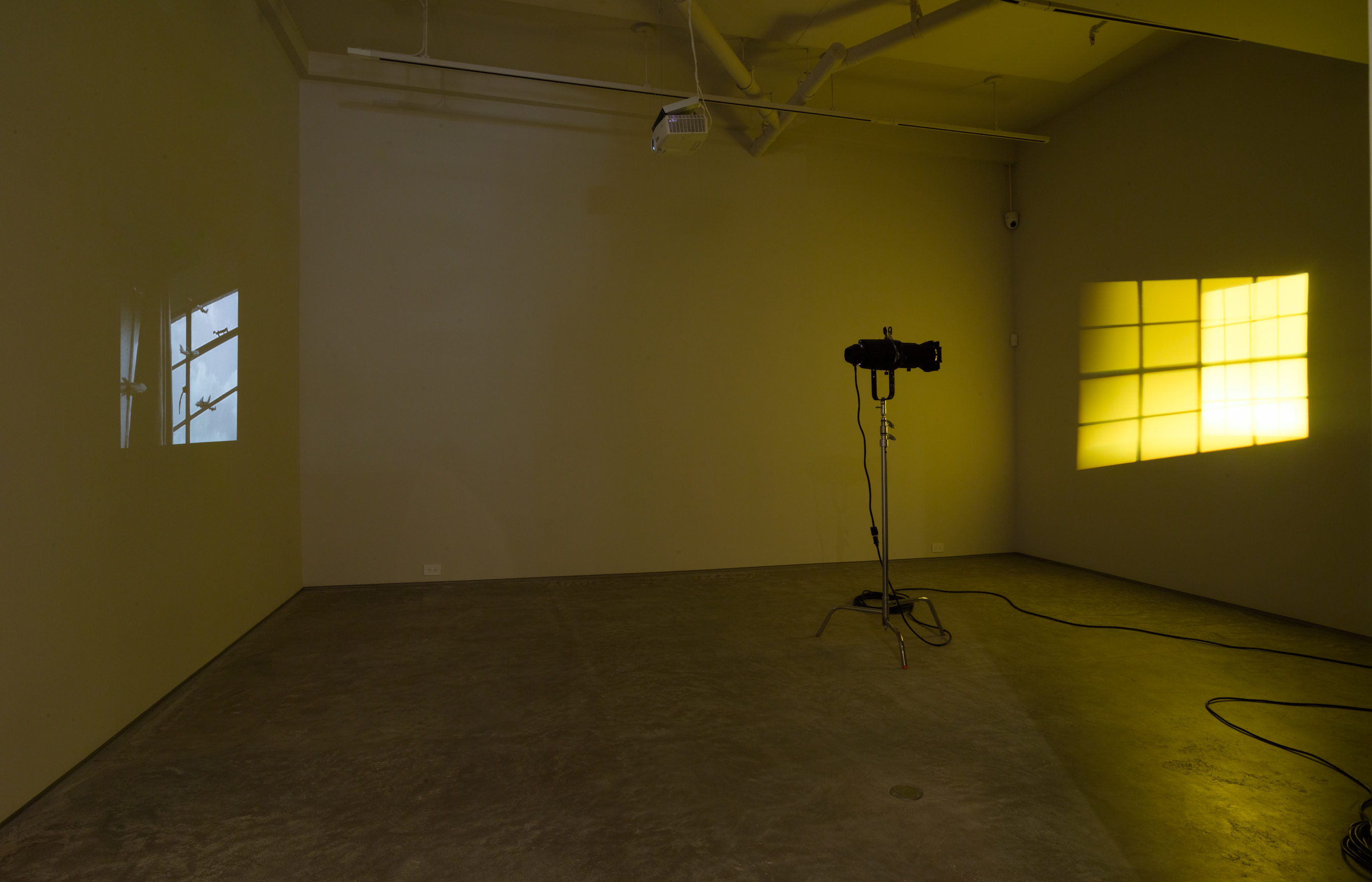 Installation shot, YOU STAND ON THE GROUND FLOOR curated by Harriet Salmon and Jesse Penridge. From left to right: Hiraki Sawa, Dwelling, 2002. Video, 9:20 minutes.  Olafur Eliasson,  Yellow Double Hung Windows, 1999. Two halogen, 60 watt profle spotlights on tripod with gobos, dimensions variable. (Credit: Jason Mandella)