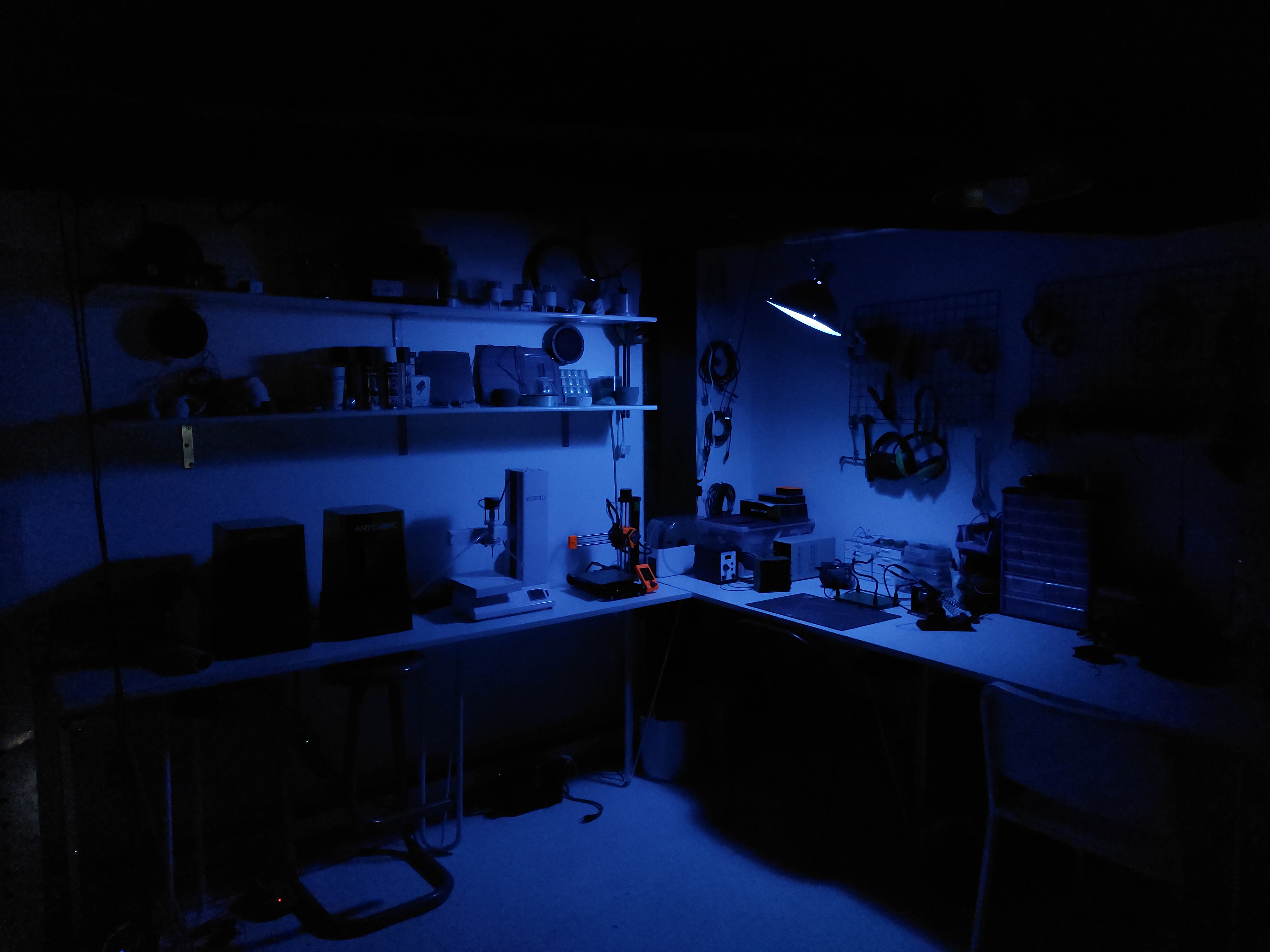 Image Description: A dark, blue-lit room that appears to be a work station. Objects are largely indiscernible in the light, but the viewer can make out shelves and a workbench with industrial equipment neatly arranged on surfaces and nailed into the wall. Below the bench, cords pool, particularly in the bottom lefthand corner of the image.