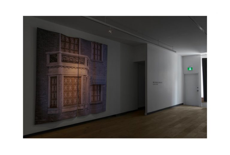 A printed photograph of a building facade juts out from the right wall as a 3D wall installation in the gallery.