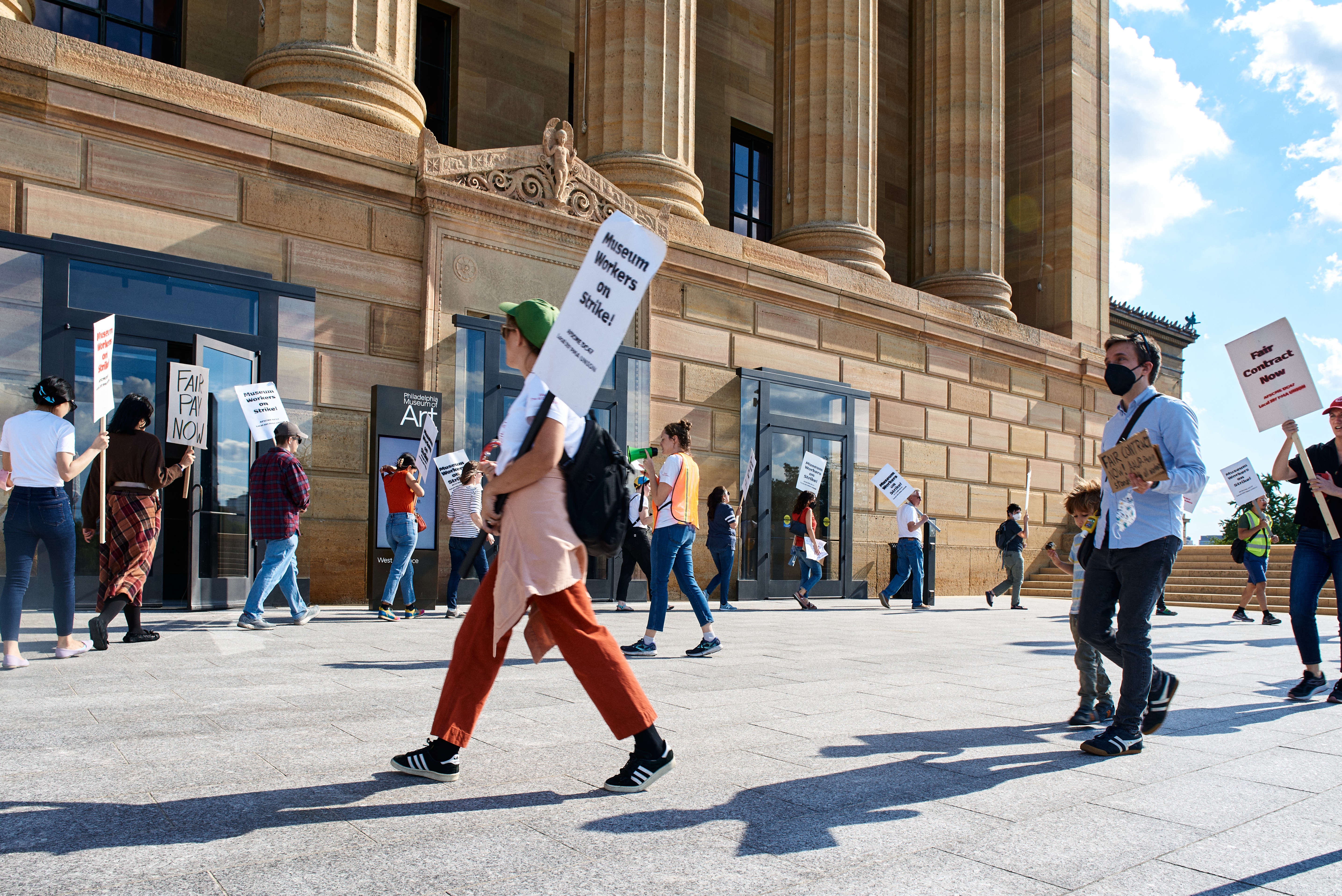 Color photo of a group of many striking workers holding picket signs in front of an entrance to the Philadelphia Museum of Art, and moving in a circle around a single worker in an orange safety vest and a small green megaphone mid-sentence, seen from slightly below.