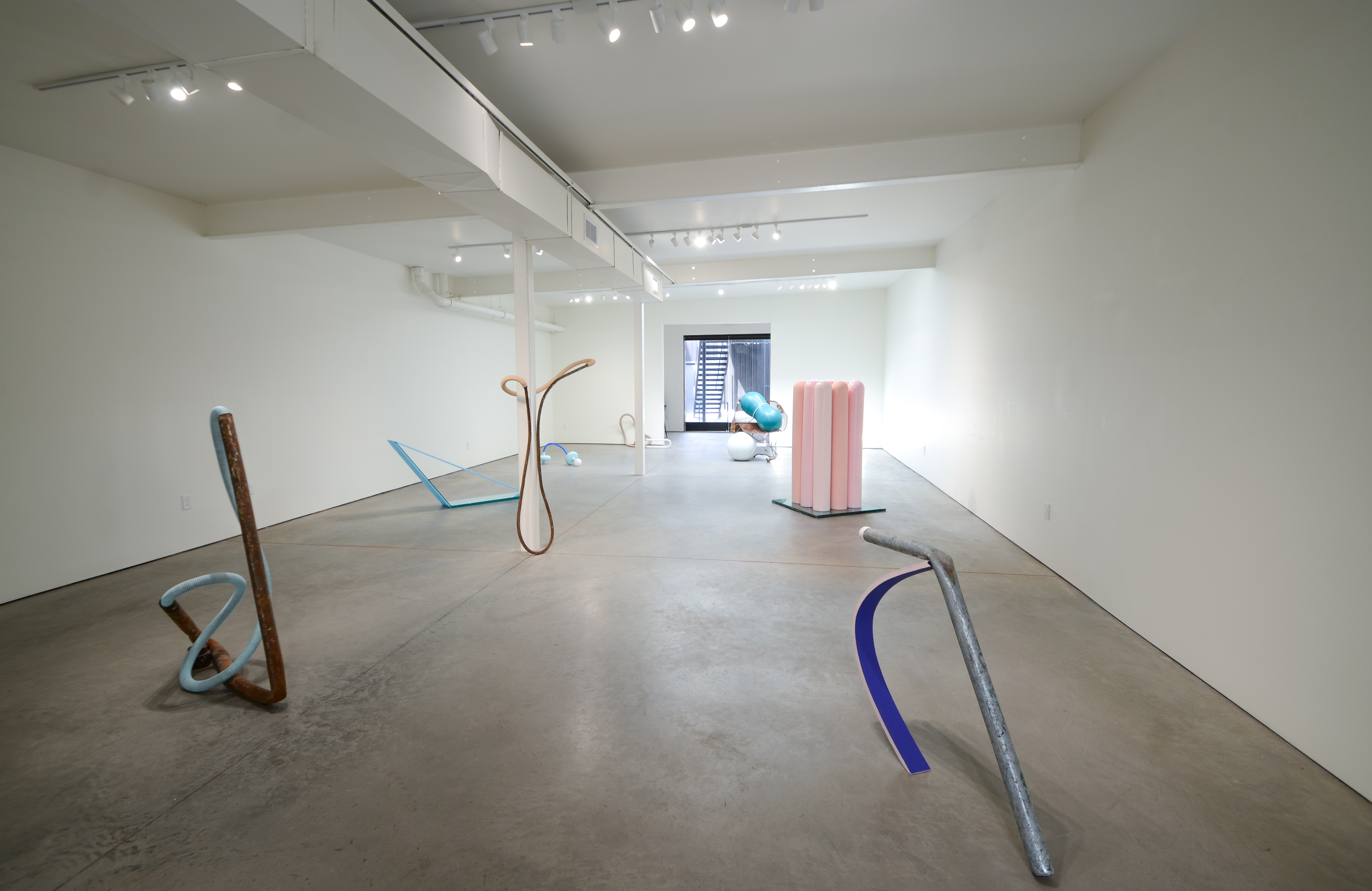 3/4’s view of a sparse white wall gallery space with concrete floors. 7-8 sculptures, roughly person-sized and evenly spaced, dot the floors of the room and are made up of various found industrial materials, heavy pipes, tubes, bent sheet metal, and so on, combined together in eccentric twists and loops. Not all, but many of the pieces are painted in bright hues of pinks, blues, and turquoises.