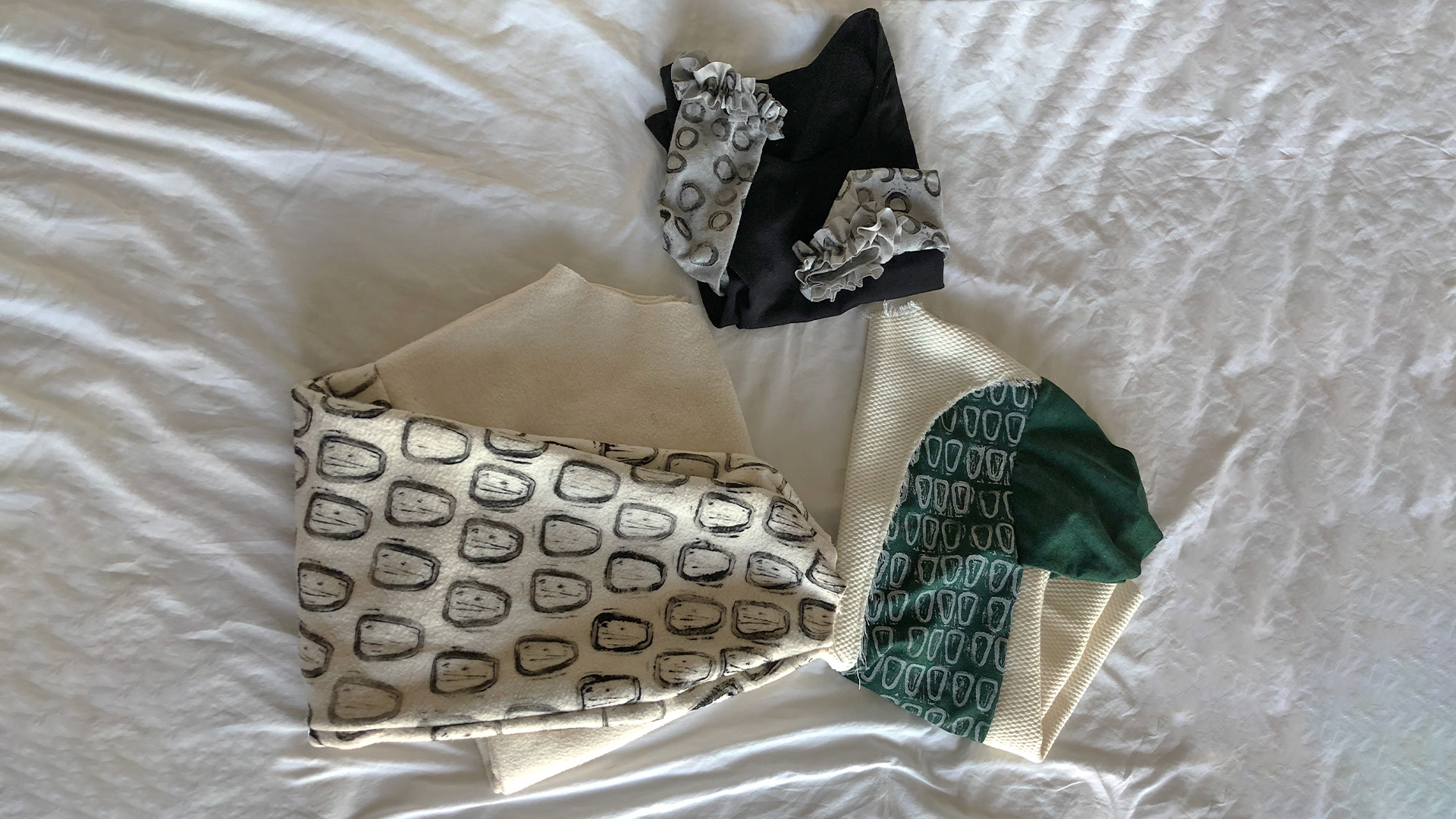 An image of three different folded clothing pieces. The one of the farthest left is a cream oversized crewneck like piece that is folded in half lengthwise. There is a black stamped icon repeating over the entirety of the sleeves. The piece in the middle is a small folded black long sleeved shirt. Midway down the sleeves there are new sewers on gray sleeves with circular black stamps repeating with ruffles circling the wrists of the sleeve. The third piece and a shirt that is made of a thick cream fabric that seems like it would be a part of a piece of furniture. The outermost parts of the shirt are dark green along with the sleeves. The green part if the body have a white repeating pattern stamped on it. These objects are placed on a wrinkly white background.