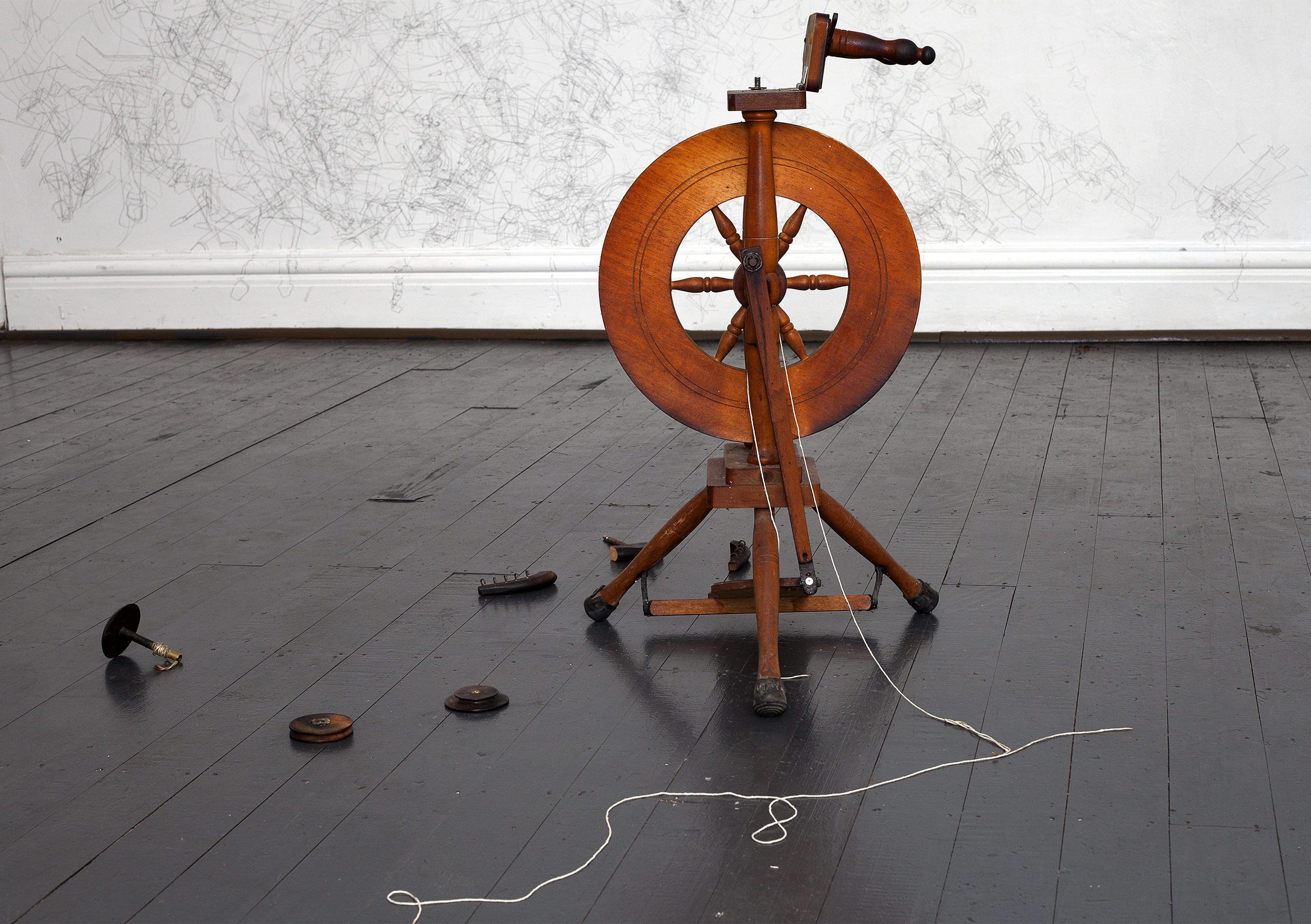 ID: A wooden spinning wheel with weaving instruments on the floor. The floor is dark wood, and the walls are painted white. The wall is covered in lead pencil marks.