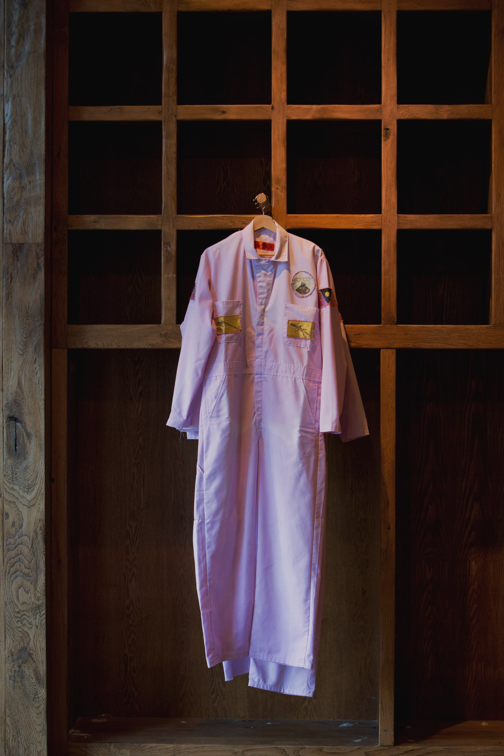 Institute of Queer Ecology Jumpsuits (2017), Felix Beaudry and Lee Pivnik for the Institute of Queer Ecology. 3 suits (S, M, L), dyed cotton and polyester blend, machine embroidered patches. Installation view of “The Symbiotic Shore” a group exhibition curated by the Institute of Queer Ecology (2021). Photo by Monica McGivern. Courtesy of the Institute of Queer Ecology, Bas Fisher Invitational and Bridge Initiative. Pink jumpsuits with embroidered patchwork displayed on a wooden shelved wall.