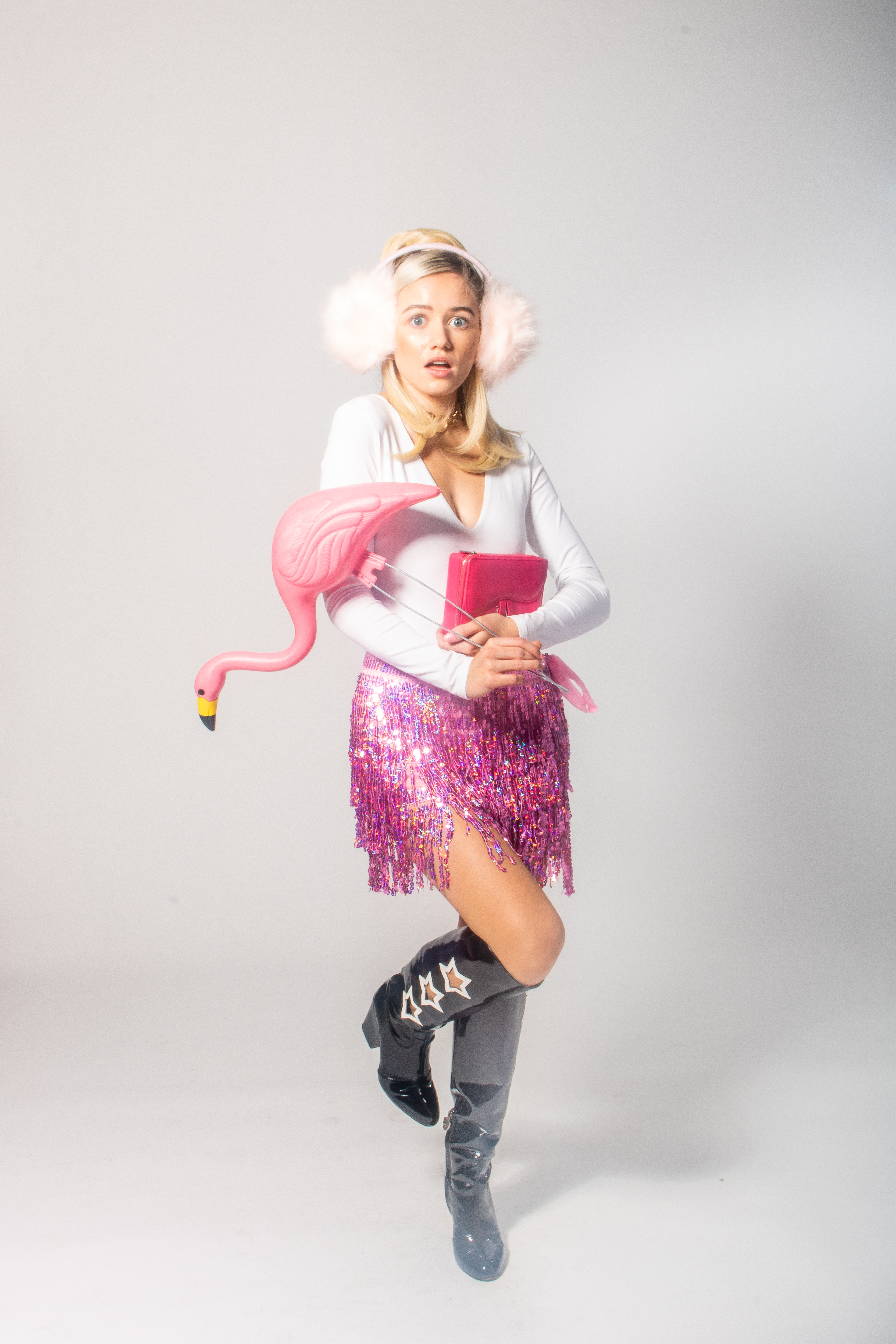 Editorial Barbie-inspired fashion portrait of blonde model Emilian Kaplan. She wears black boots, a pink glitter skirt, a white long-sleeve top, pink earmuffs, and holds a pink flamingo and book—an eclectic, chaotic display mirroring Barbie’s ‘everything’ lifestyle. Shot against a crisp white backdrop.