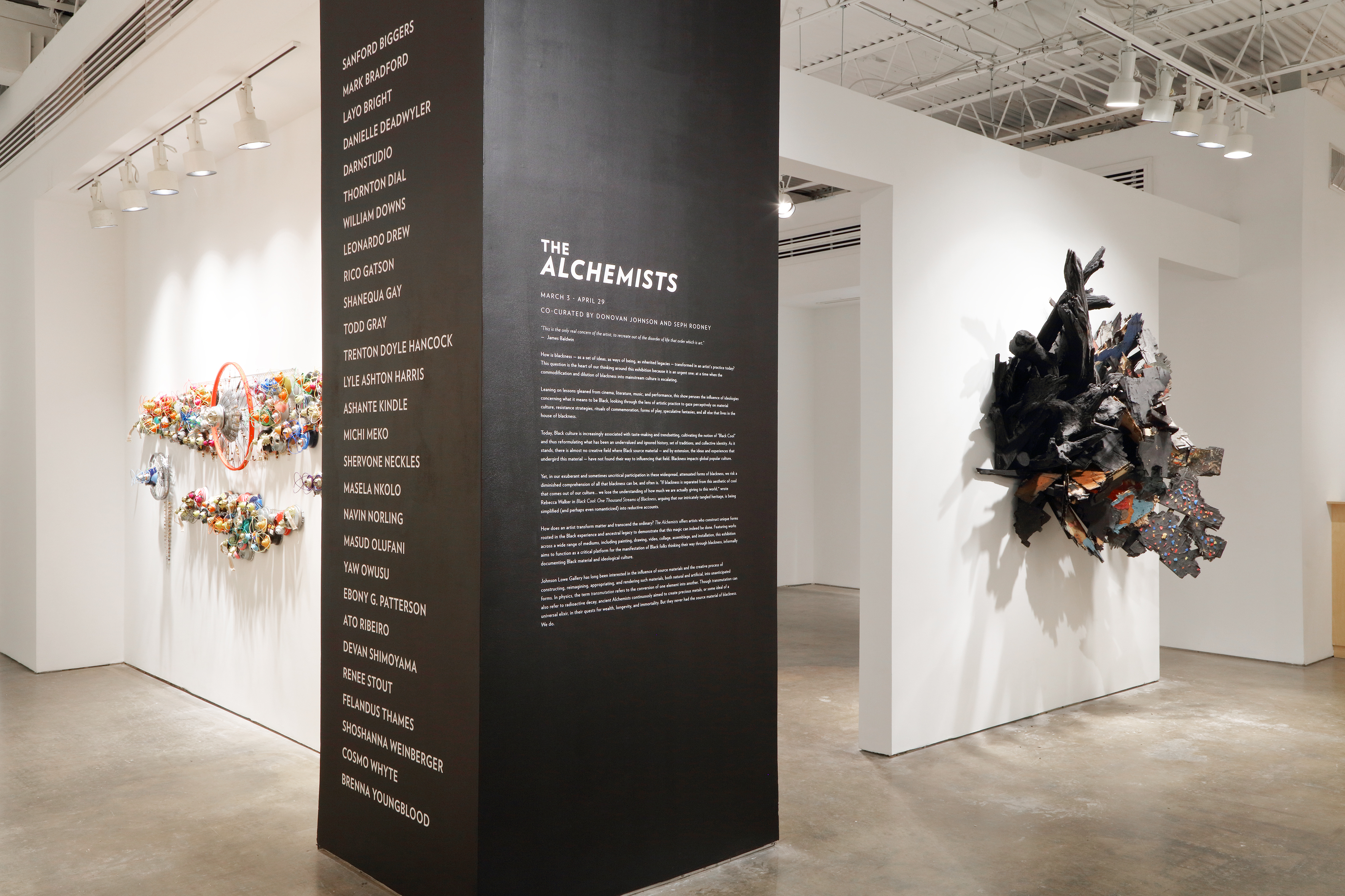 Art exhibition in a white wall gallery space. One section of a corner has been painted black and has white lettering that reads the exhibition title: “The Alchemists.”