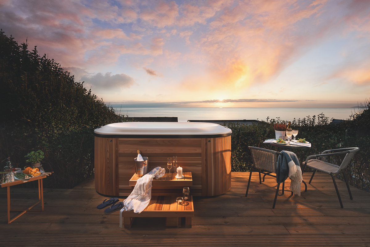 Timber patio area with jacuzzi, bottle in cooler, and outdoor furniture. Sea and sunset in background.