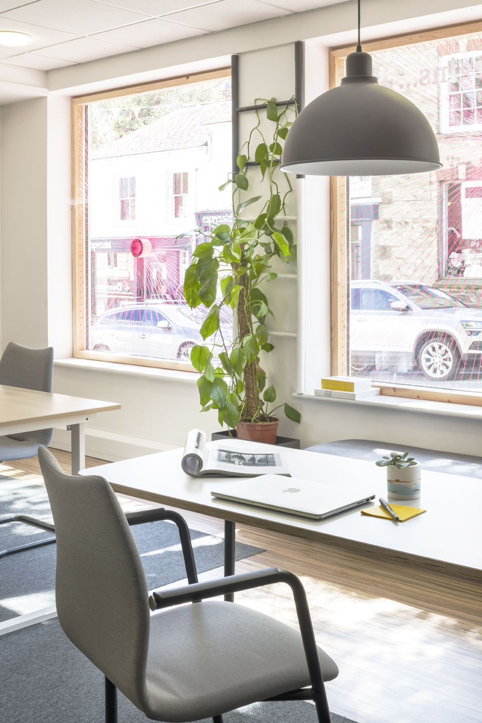 Close-up of desk with book, laptop and pot plant. Large grey ceiling pendant suspended above. Pot plant and windows in background.