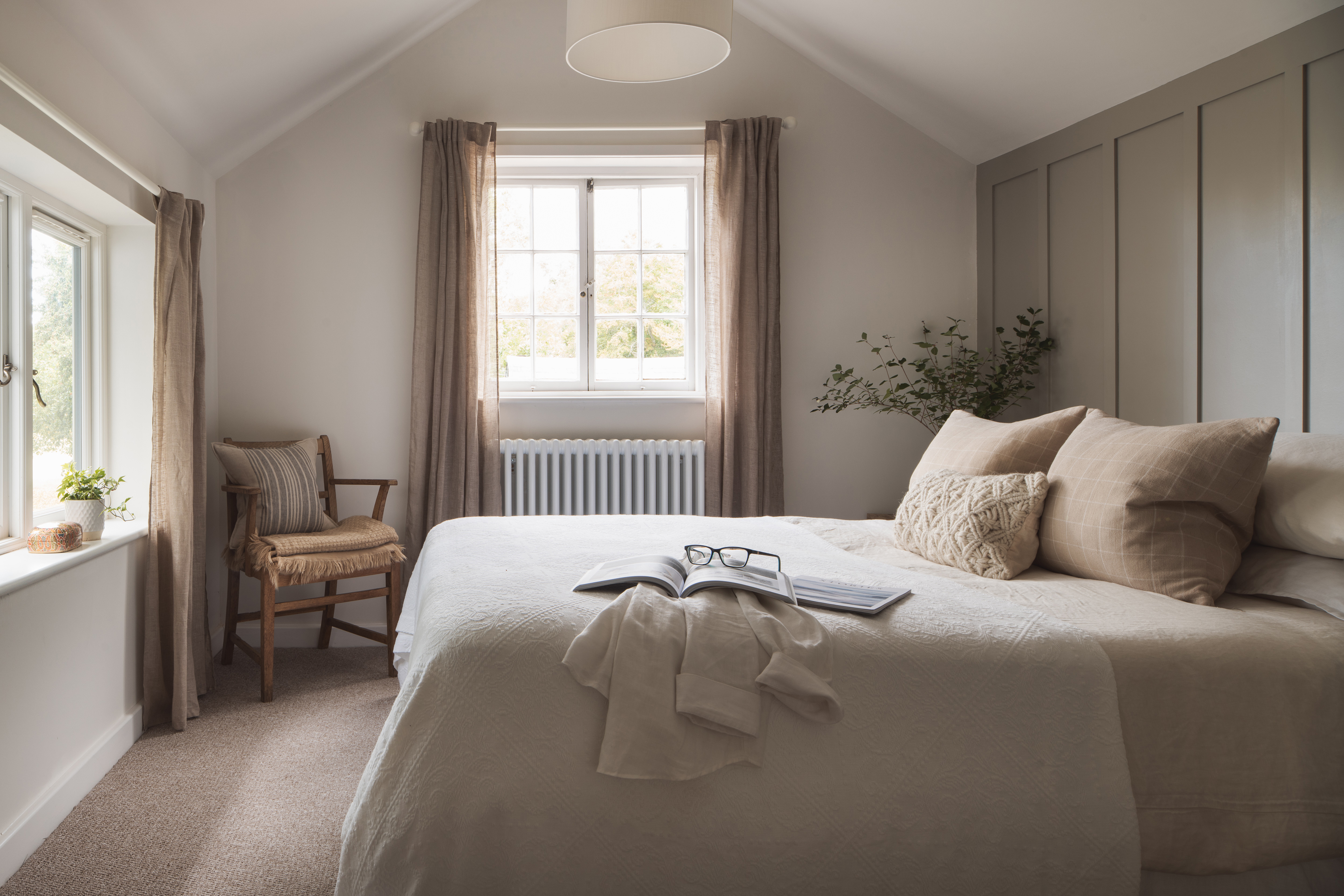 Bedroom with double bed in neutral colour palette. Panels on one wall, two windows with long curtains and chair in corner.