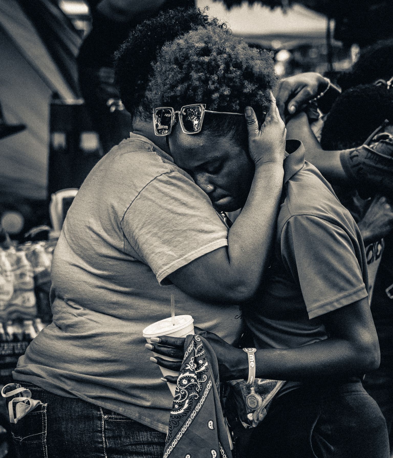A black and white photograph with high contrast shows two people embracing, seemingly, in mourning. They both wear t-shirts and jeans, one is holding a bandana and a takeout drink from a restaurant.
