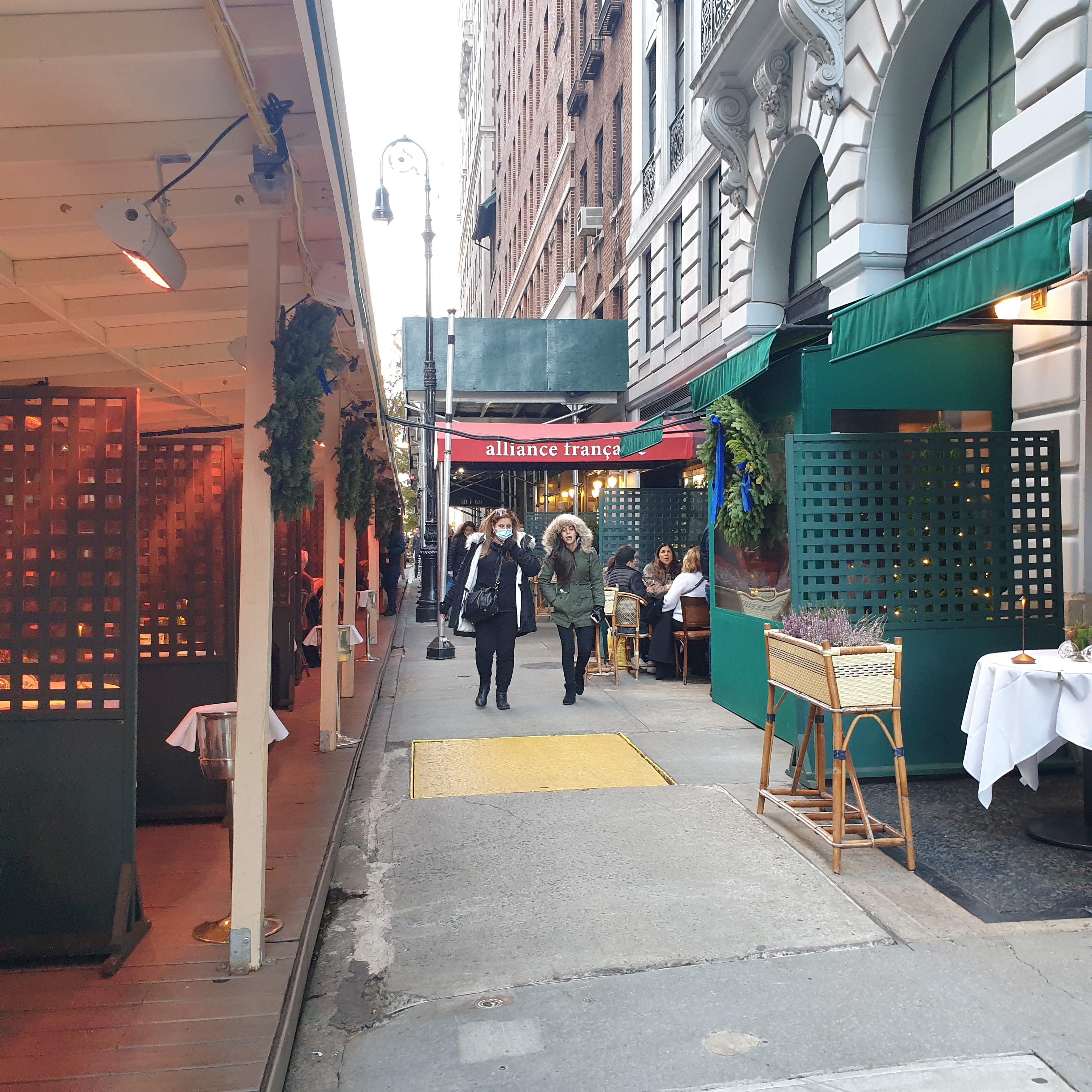 A sidewalk in New York. To the left is an enclave with seating for dining, to the right storefronts with green planters and awnings. Two women can be seen walking on the sidewalk. Behind them is a read awning reading alliance française.